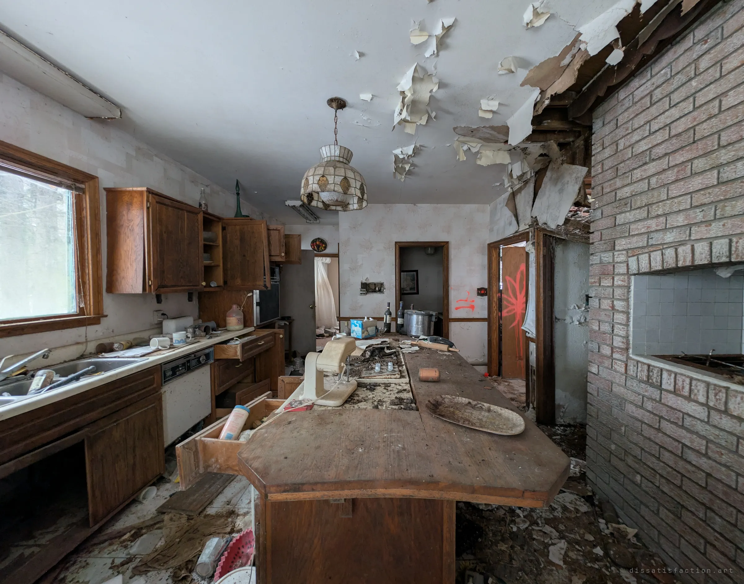 A long wooden kitchen island still set with a mixer, a platter and bottles, while the ceiling paper curls down in sheets. A spray-painted plant marks a doorway beyond.