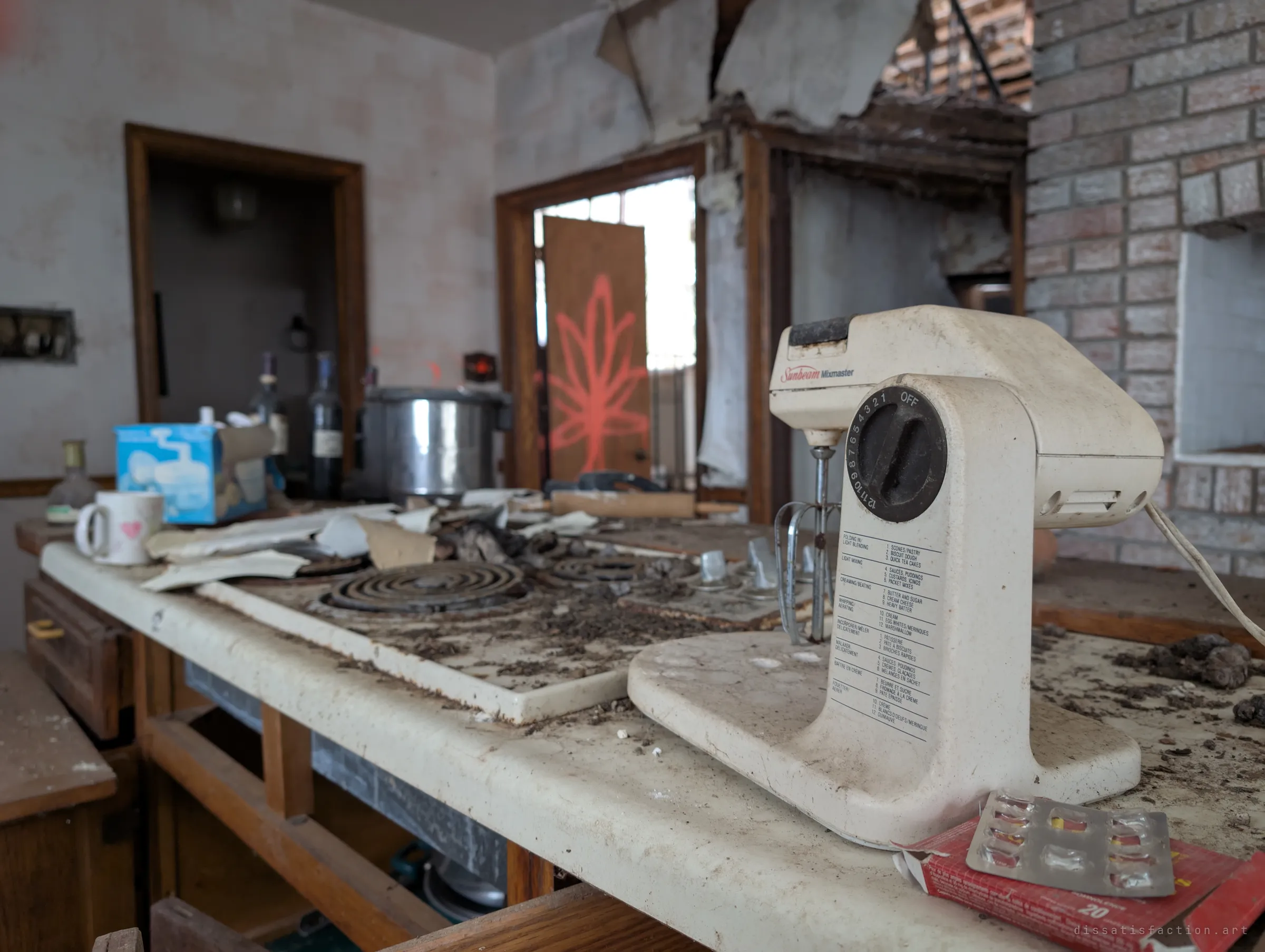 Close-up of a dusty stand mixer on the stovetop counter, next to an opened blister pack, with the graffiti-marked doorway blurred behind. These small objects make the house feel abandoned in the middle of an ordinary day.