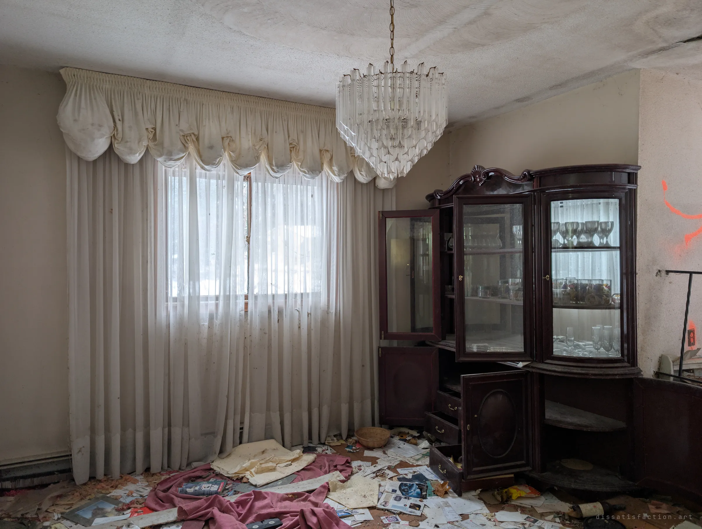A formal dining room with ruffled valance curtains, a crystal chandelier and a glass-fronted china cabinet still holding wine glasses. Magazines, mail and a pink blanket cover the floor.