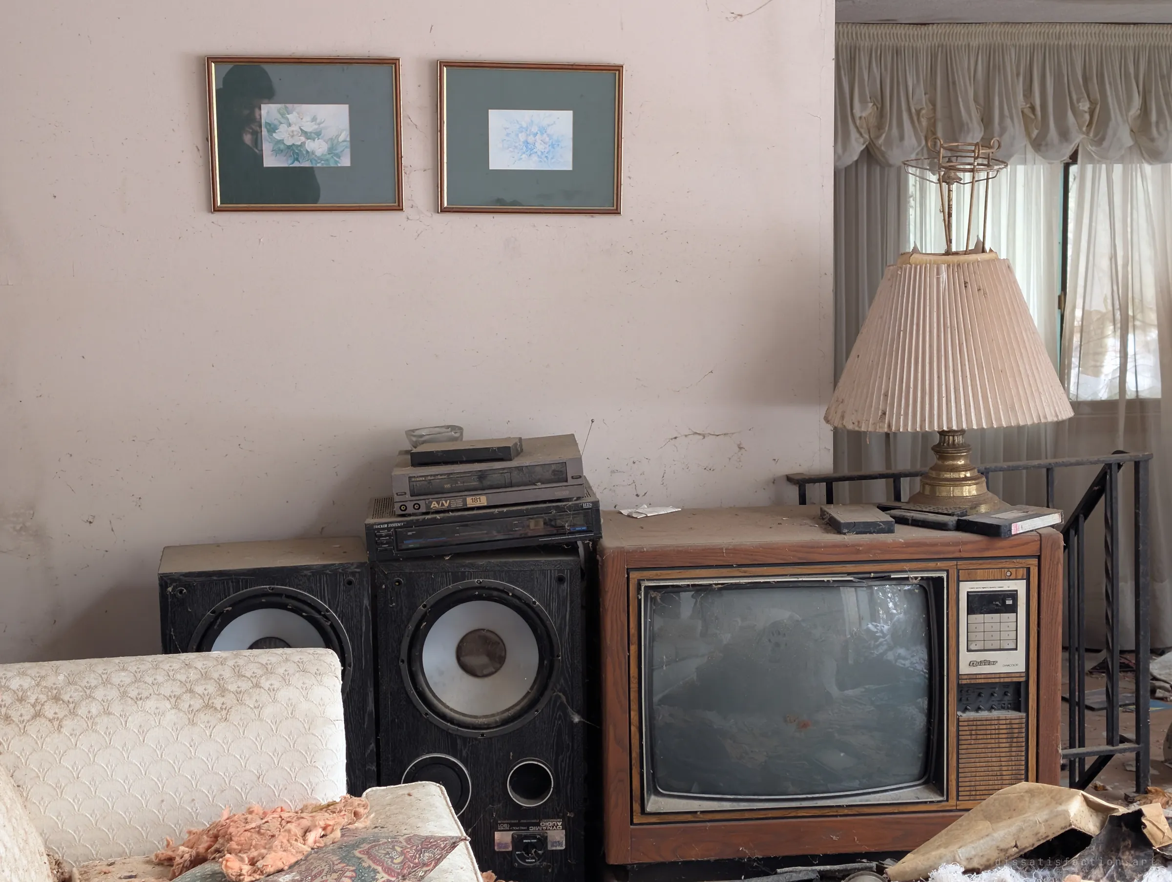 A dusty living-room corner: two floral prints above stacked VCRs, big floor speakers and a wood-cabinet tube TV, with a pleated lamp by a curtained window. The whole entertainment setup was left exactly as it stood.