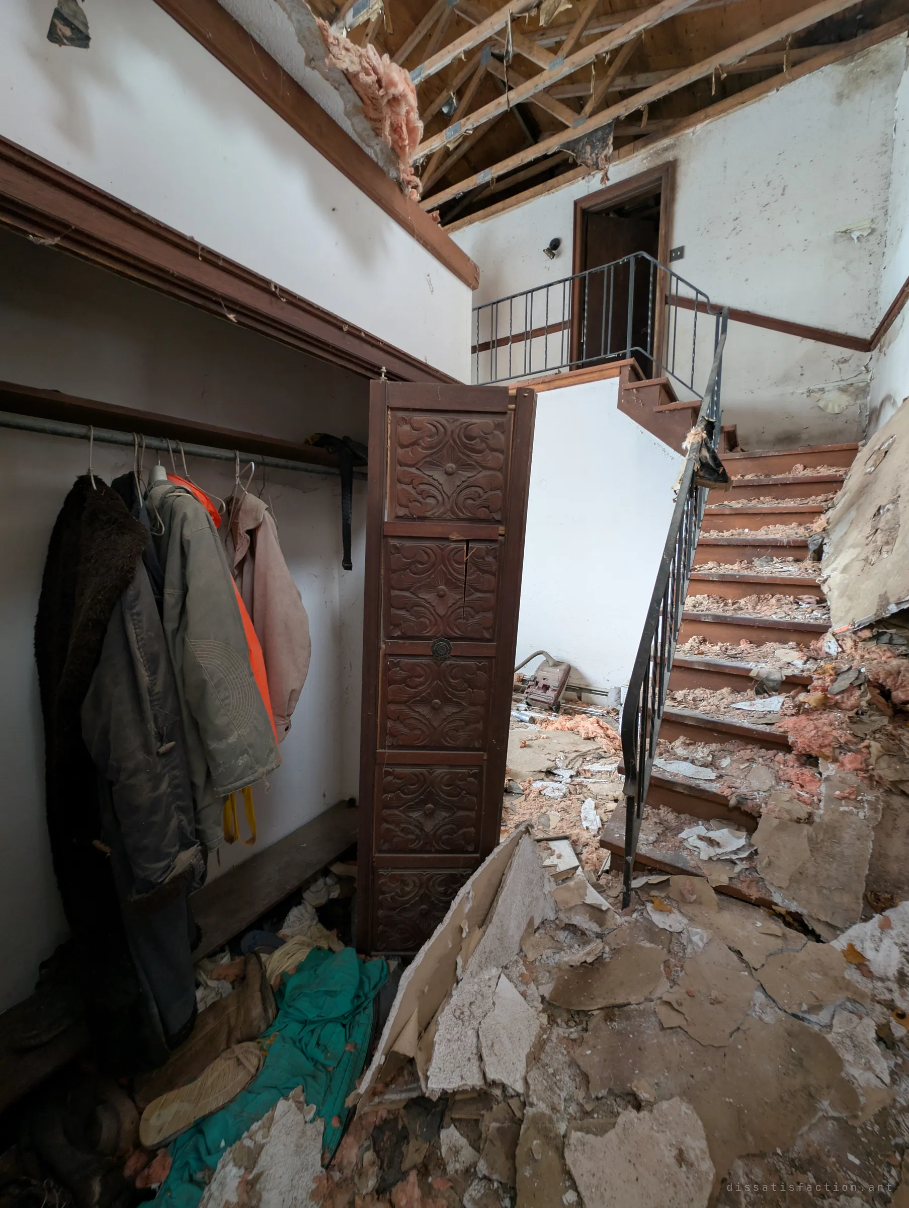 An entry hall with coats still hanging in an open closet beside a carved wooden door, while a stair littered with insulation climbs under a ceiling torn open to the rafters. Everyday belongings sit next to the collapse.