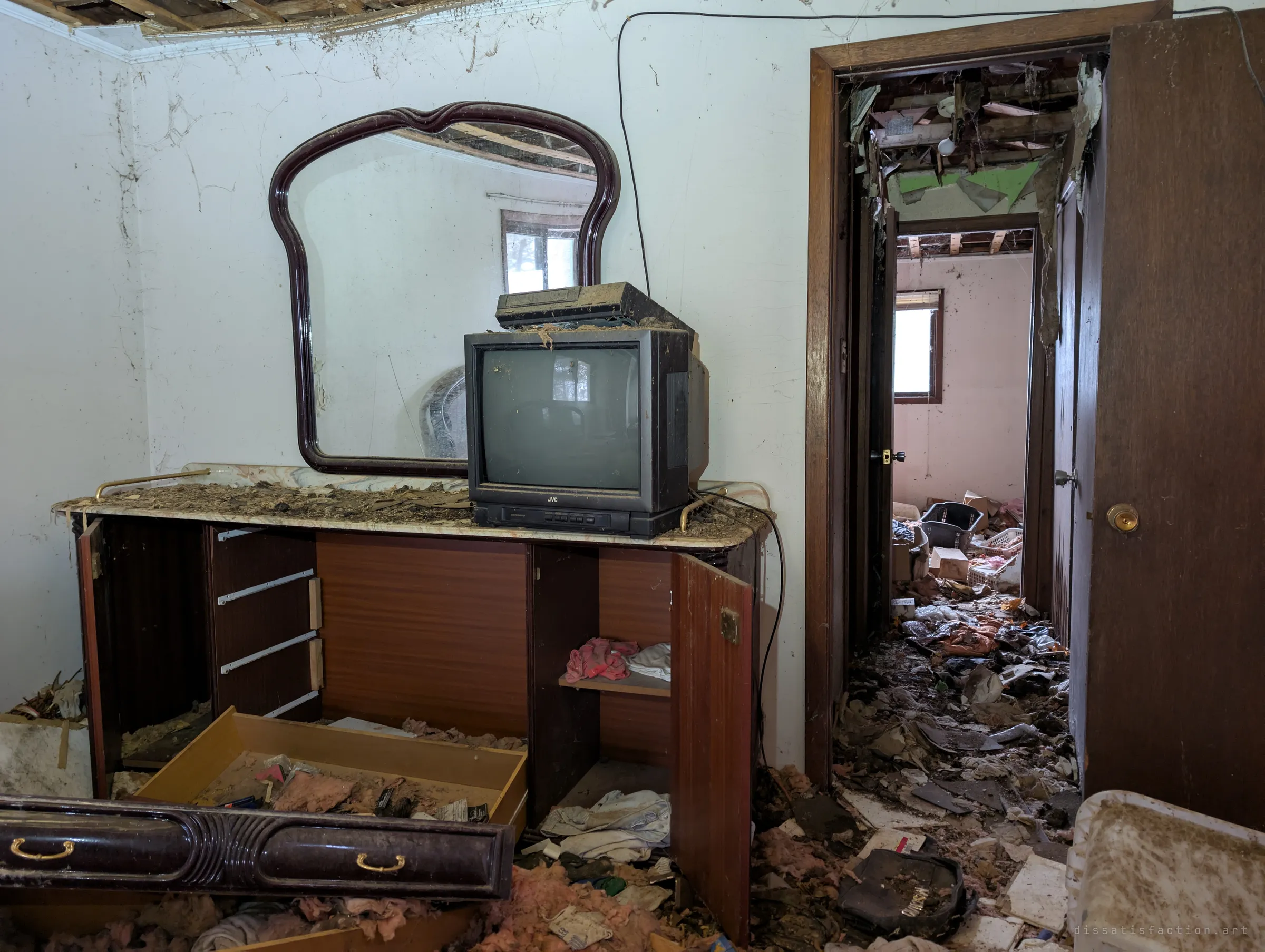 A bedroom dresser with a scalloped mirror and a small tube TV, drawers pulled out, looking through a doorway down a hall buried in debris. The open ceiling above the hall shows how far the decay reaches.