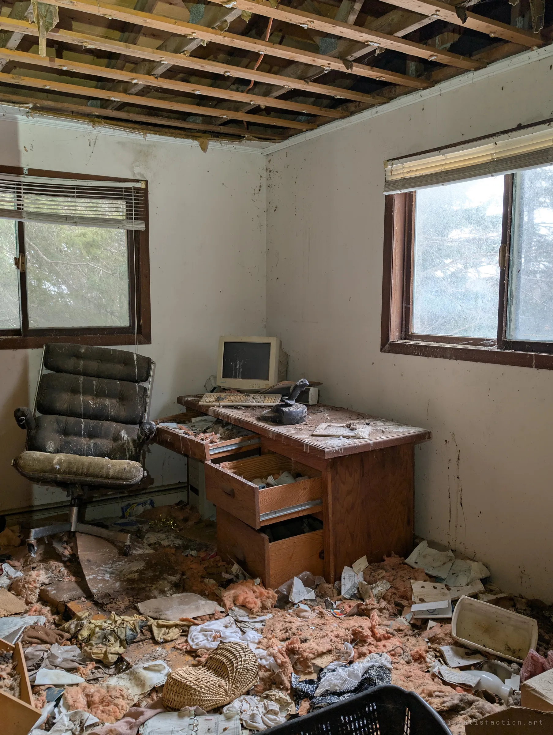 A small home office with a beige CRT monitor and keyboard on a wooden desk, a tufted office chair beside it and exposed ceiling joists overhead. Insulation and papers cover the floor.
