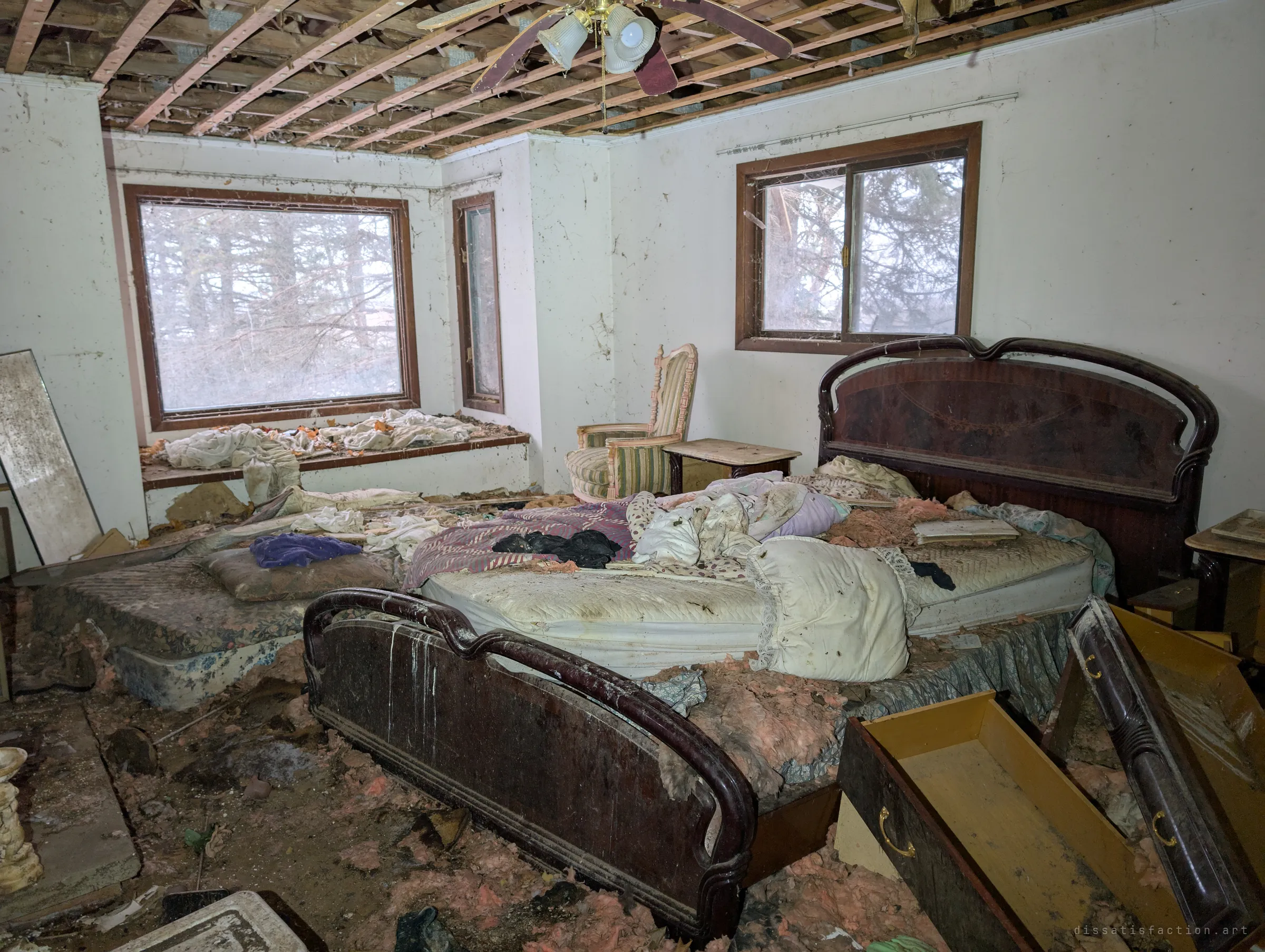 A main bedroom with a glossy dark headboard and footboard around a stripped, dirt-covered mattress, pulled-out drawers and a striped armchair by the windows. Bare ceiling joists and a dangling fan hang overhead.