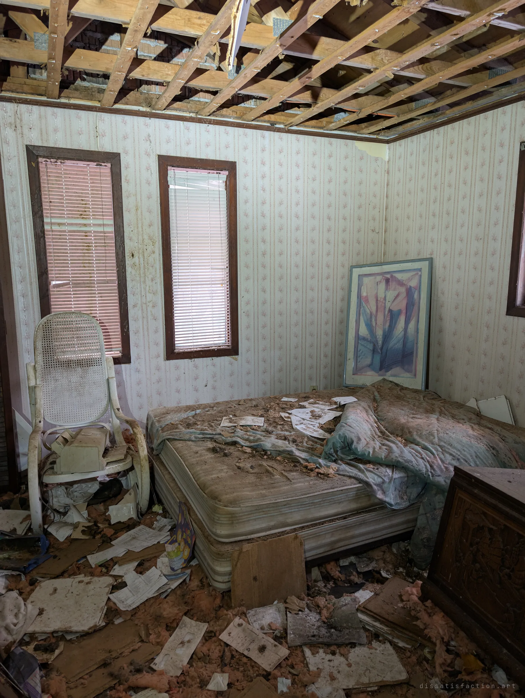 A smaller bedroom with floral wallpaper, a cane rocking chair and a bare mattress scattered with papers under an open ceiling. A framed abstract print still leans in the corner.