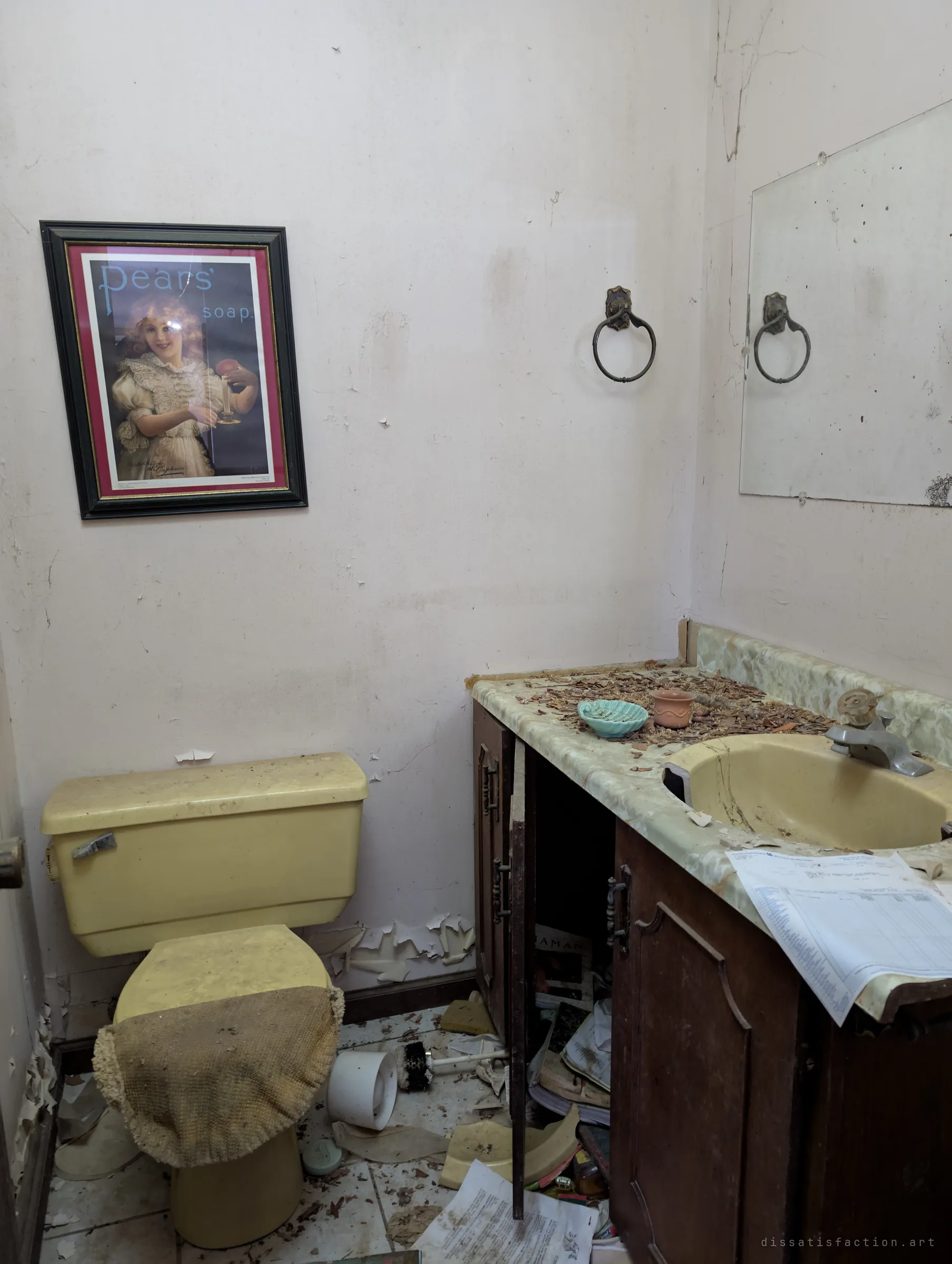A powder room with a yellow toilet and sink, a vintage soap-advert print on the wall and papers spilling from the vanity. The period fixtures date the house.