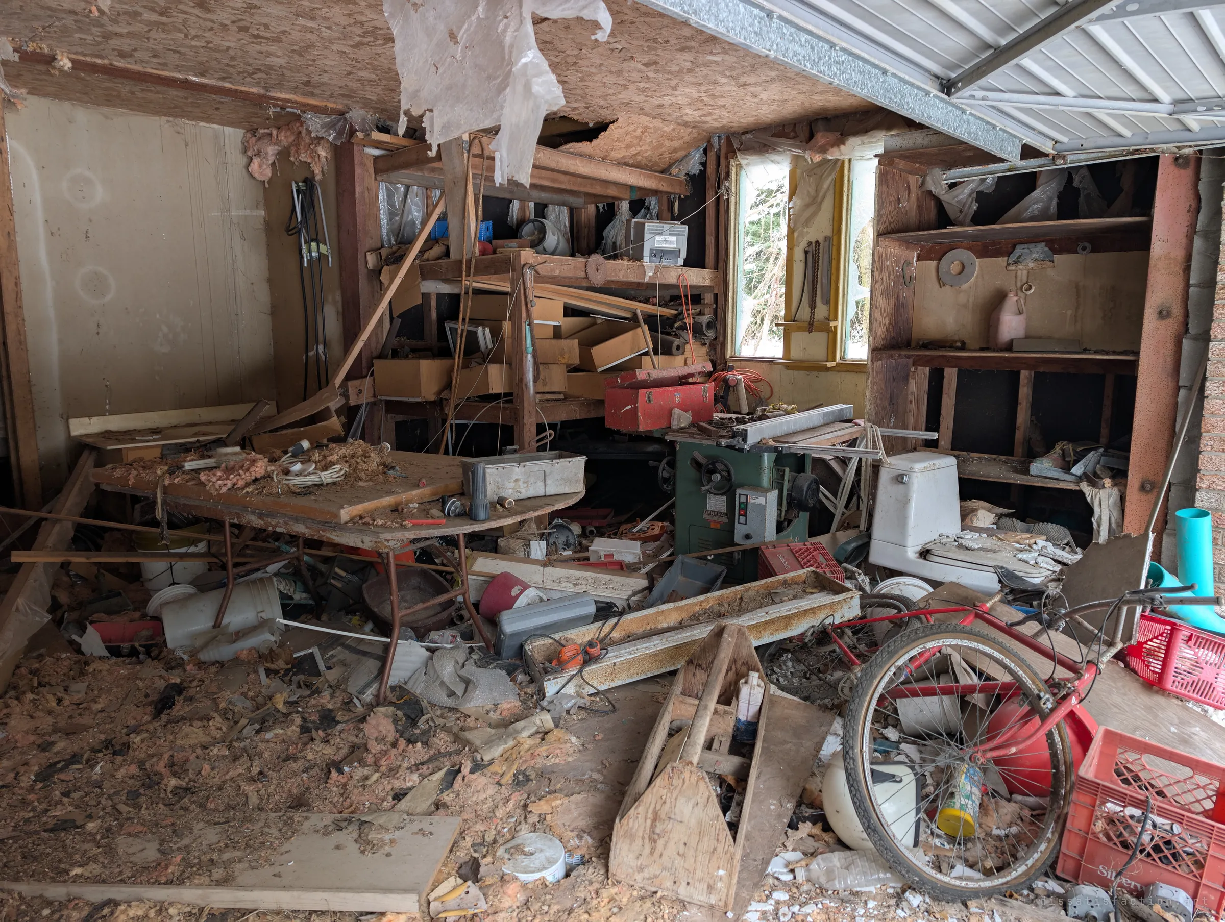 A garage workshop crammed with a table saw, a toolbox, shelving and a red bicycle, beneath a half-open overhead door and a sagging ceiling. Decades of tools and clutter remain in place.
