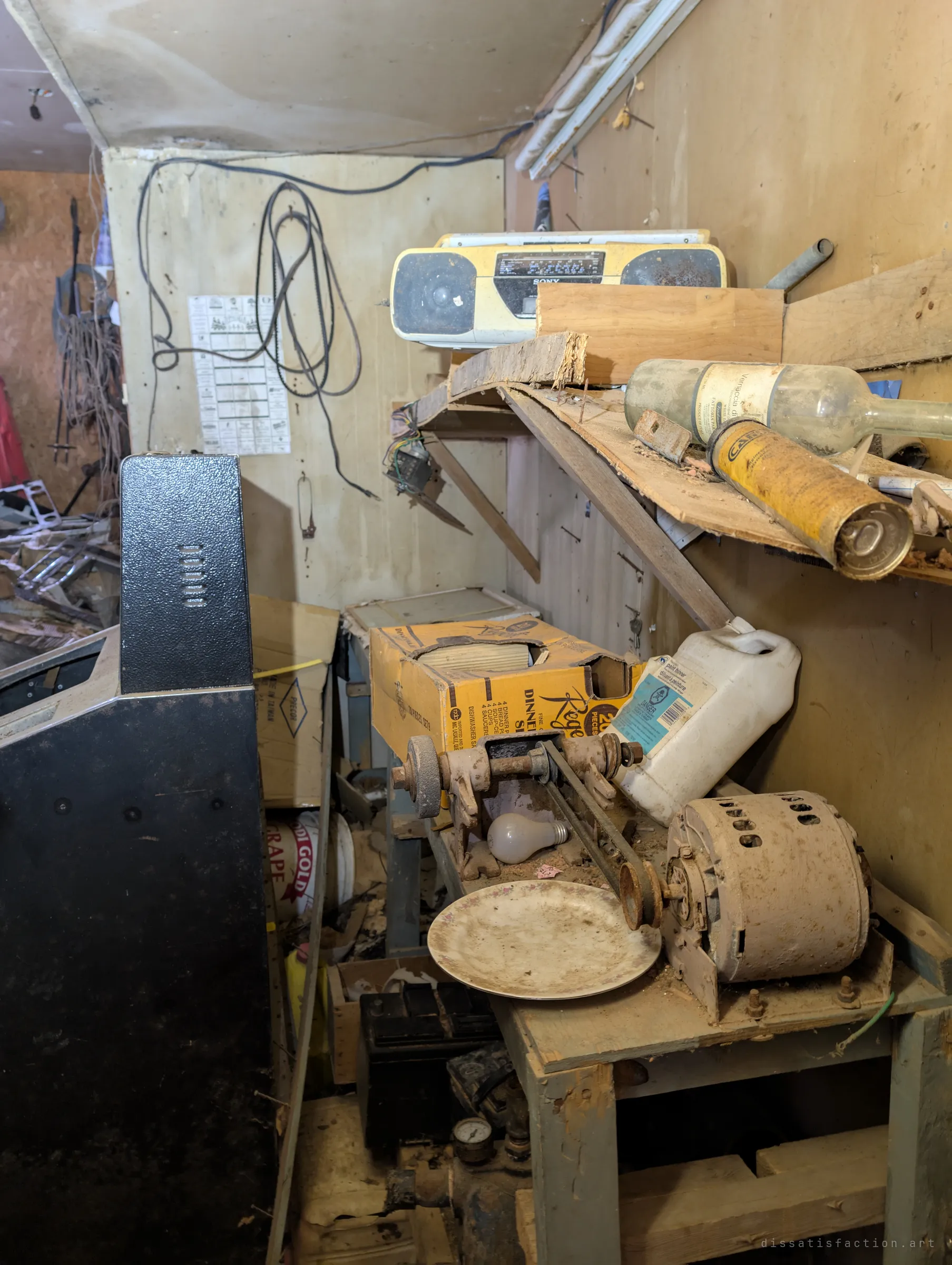 A cramped workbench corner with a yellow boombox on a broken shelf, an old bench grinder and electric motor, and a tall black cabinet in the foreground. Belts and cables hang on the wall behind.