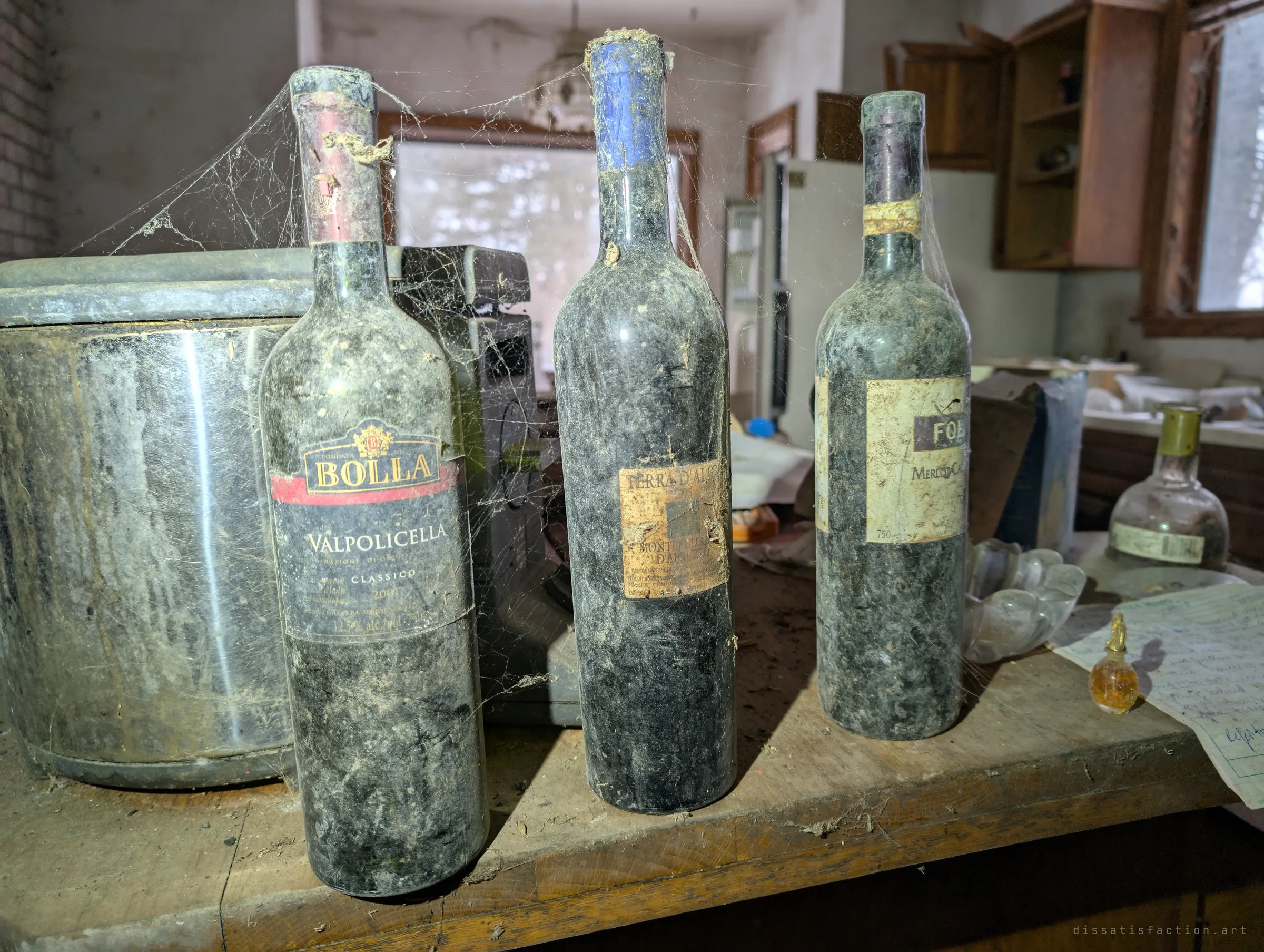 Three cobweb-draped wine bottles from around 2000 stand on a dusty kitchen counter next to a steel pot, with the abandoned kitchen blurred behind. Spider silk ties the bottles together, a measure of how long nobody has touched them.