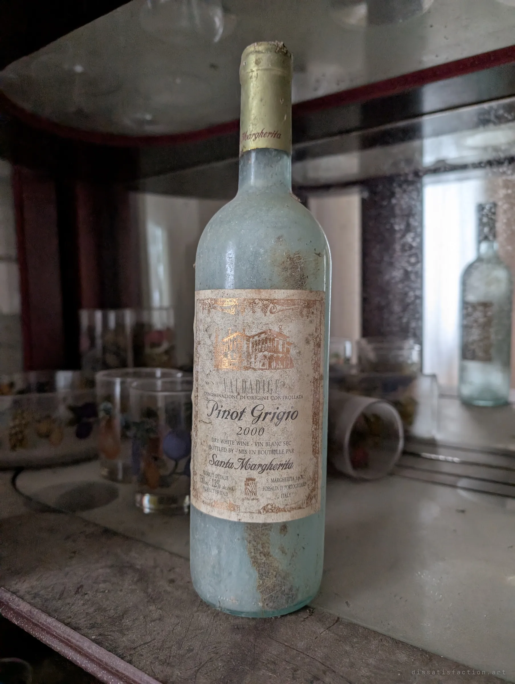 A frosted, dust-caked bottle of 2000 pinot grigio stands alone on a mirrored cabinet shelf among painted fruit glasses. The unopened bottle hints at an occasion that never came.