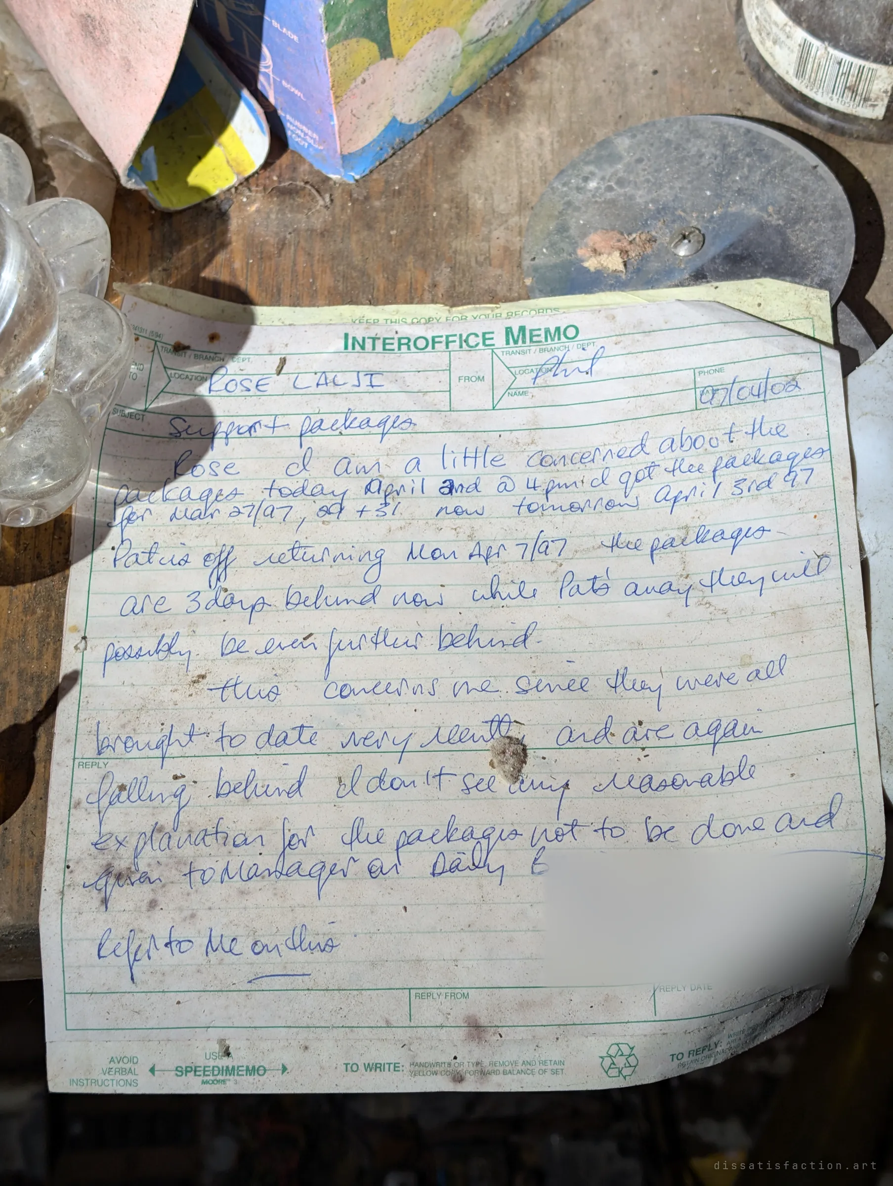 A handwritten 1997 interoffice memo complaining about late packages lies on a dusty workbench beside a glass dish and tools. A stray piece of someone’s working life, kept for decades in the house.