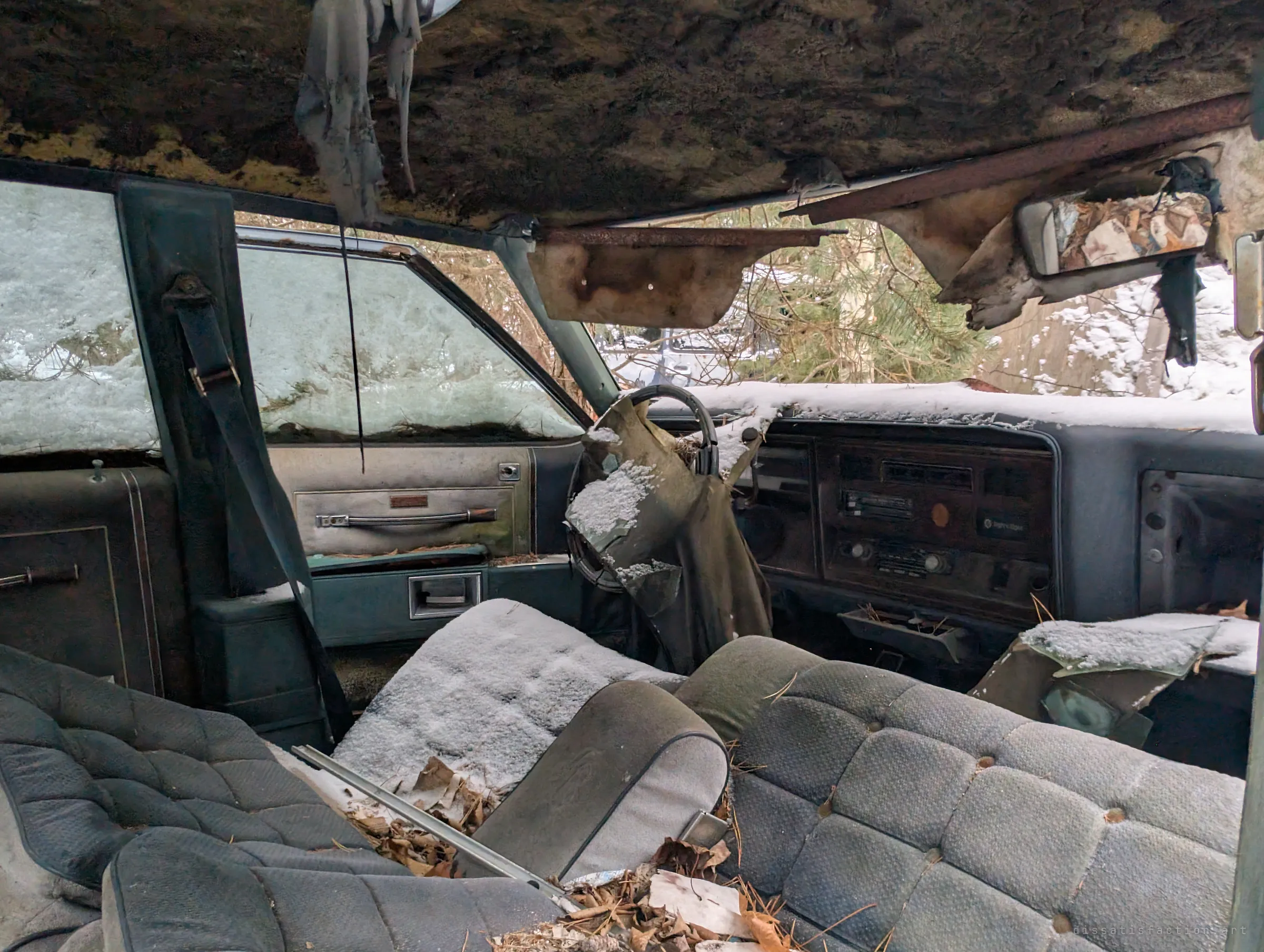 Inside an older car with a bench seat, snow settles on the upholstery, the steering wheel is wrapped in rotting fabric and the headliner hangs in shreds. Pine branches press against the frosted windows.