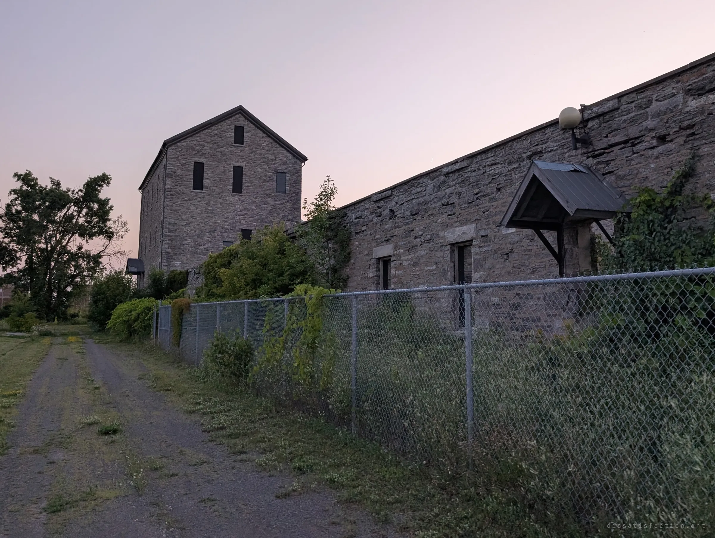 A tall gabled stone mill tower rises at dusk behind a long, vine-covered stone wing and a chain-link fence, with an overgrown gravel track leading along it. Sets the scene: a fenced, half-forgotten stone industrial complex.