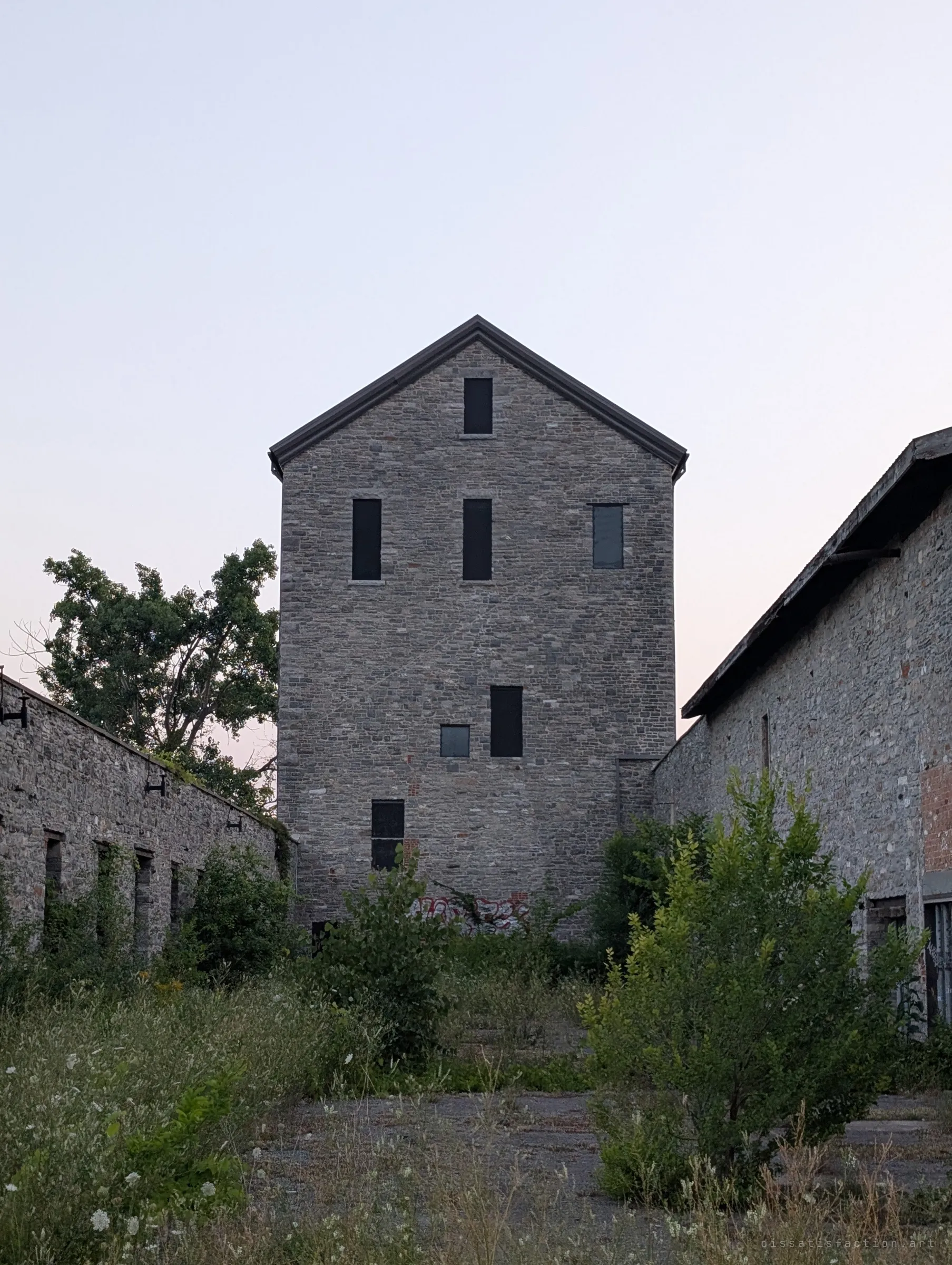 The stone mill tower seen head-on down an overgrown courtyard between two roofless stone wings, its dark window openings scattered up the facade. Shows the scale of the tower against the ruins around it.