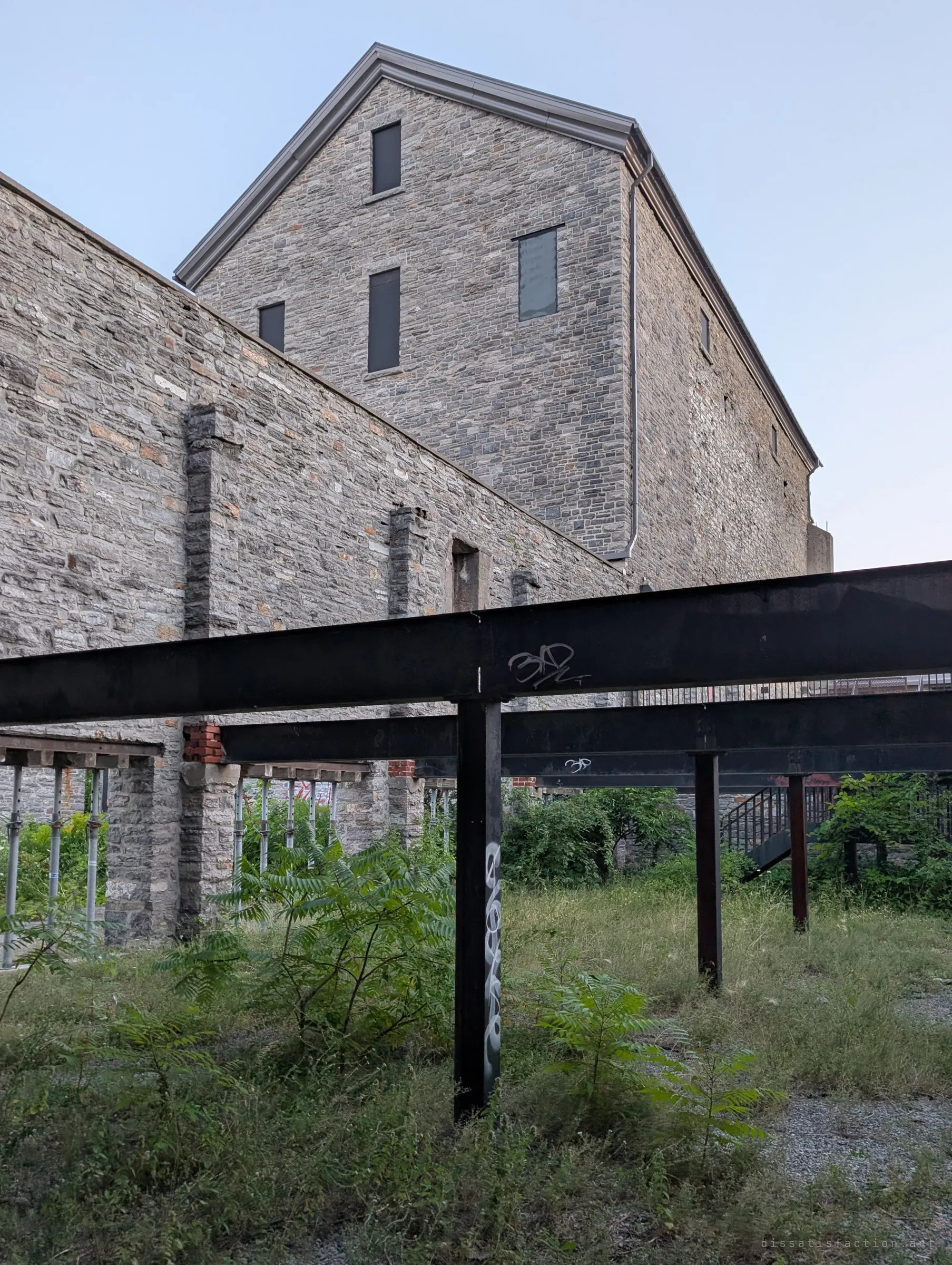 Looking up past rusted steel I-beams on posts, tagged with graffiti, to a stone wall and the gabled mill tower against a pale sky. The steel frame props up what is left of the old walls.