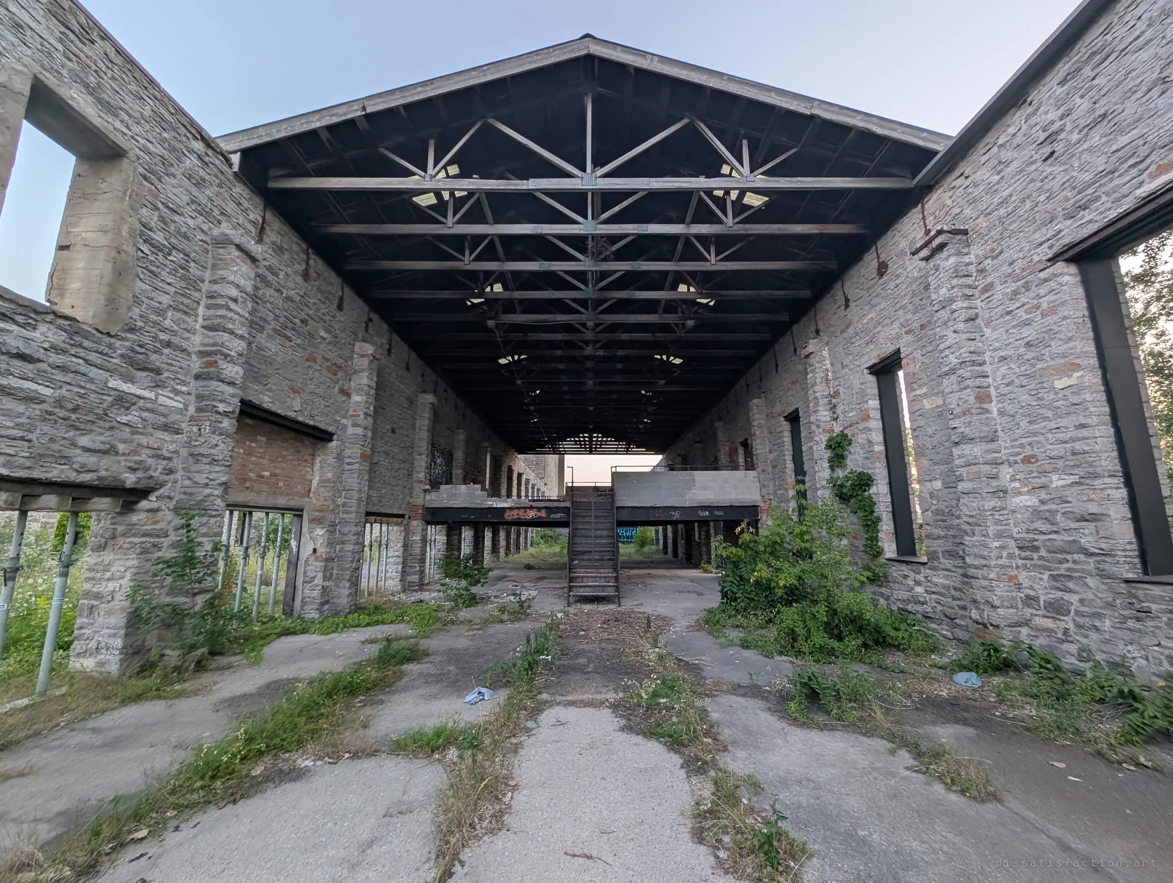A long roofless stone hall under an open timber truss canopy, cracked concrete floor sprouting weeds and a metal stair leading up to a raised platform at the far end. The ultra-wide view shows the whole gutted shell of the mill.
