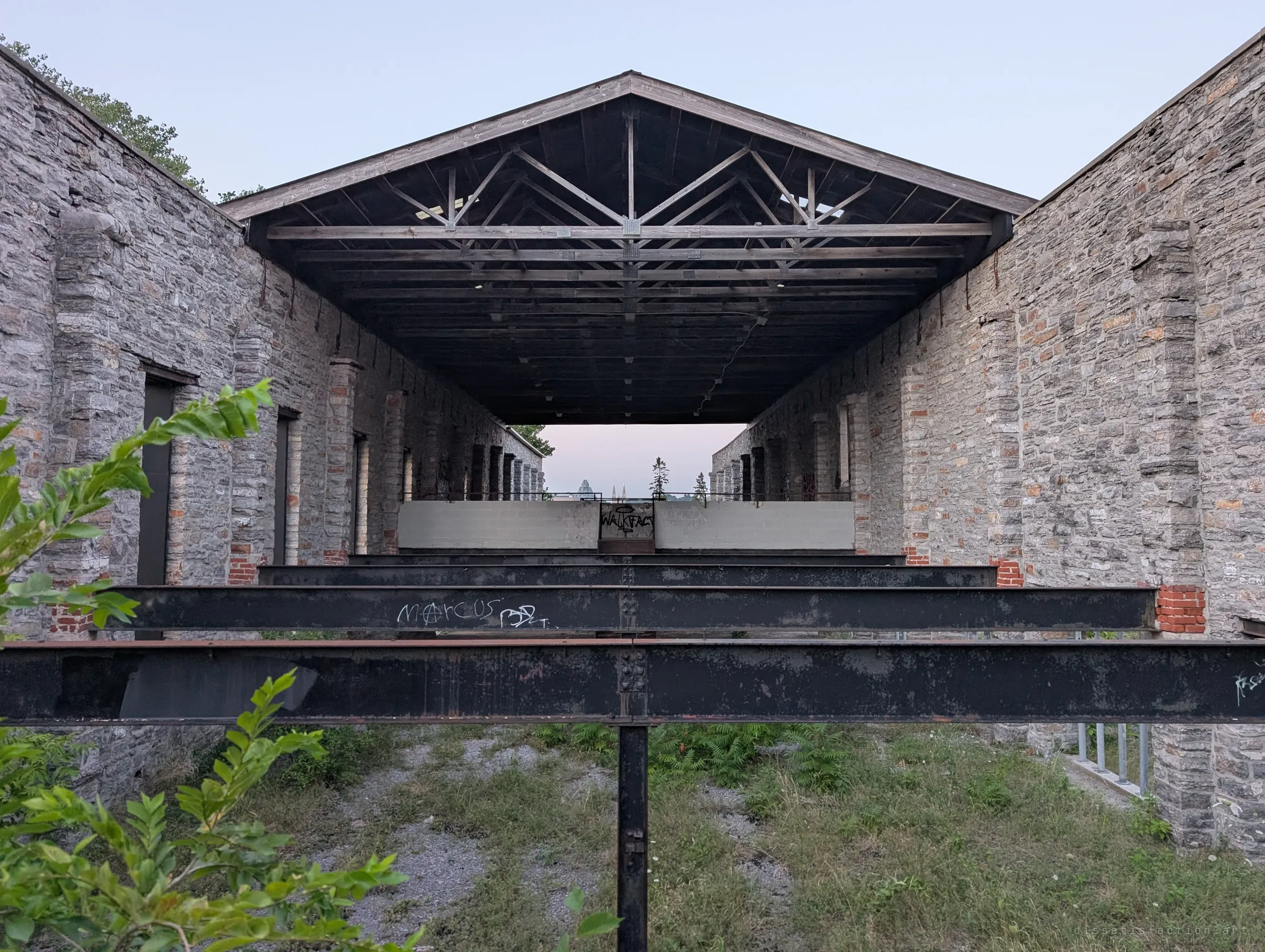 Rusted steel beams stacked in perspective beneath a timber truss roof, framed by two stone walls, leading the eye to a bright opening at the far end. The layered beams and trusses make a tunnel-like symmetry.