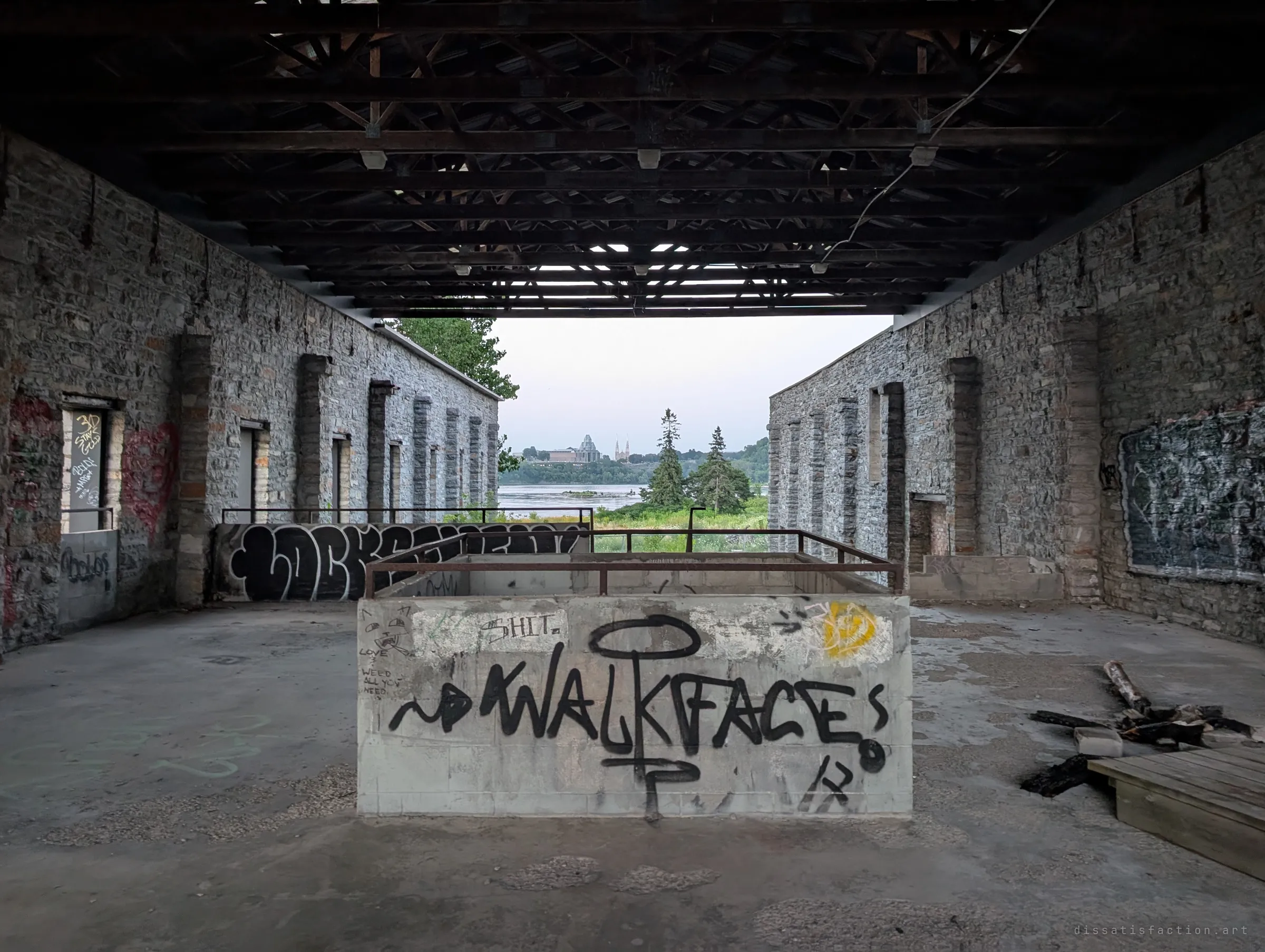 Under a dark steel-and-timber roof, a graffiti-covered concrete block sits in the middle of a roofless stone hall, with the far end opening onto trees, a river and a distant skyline. The view out shows how close the ruin sits to the water.