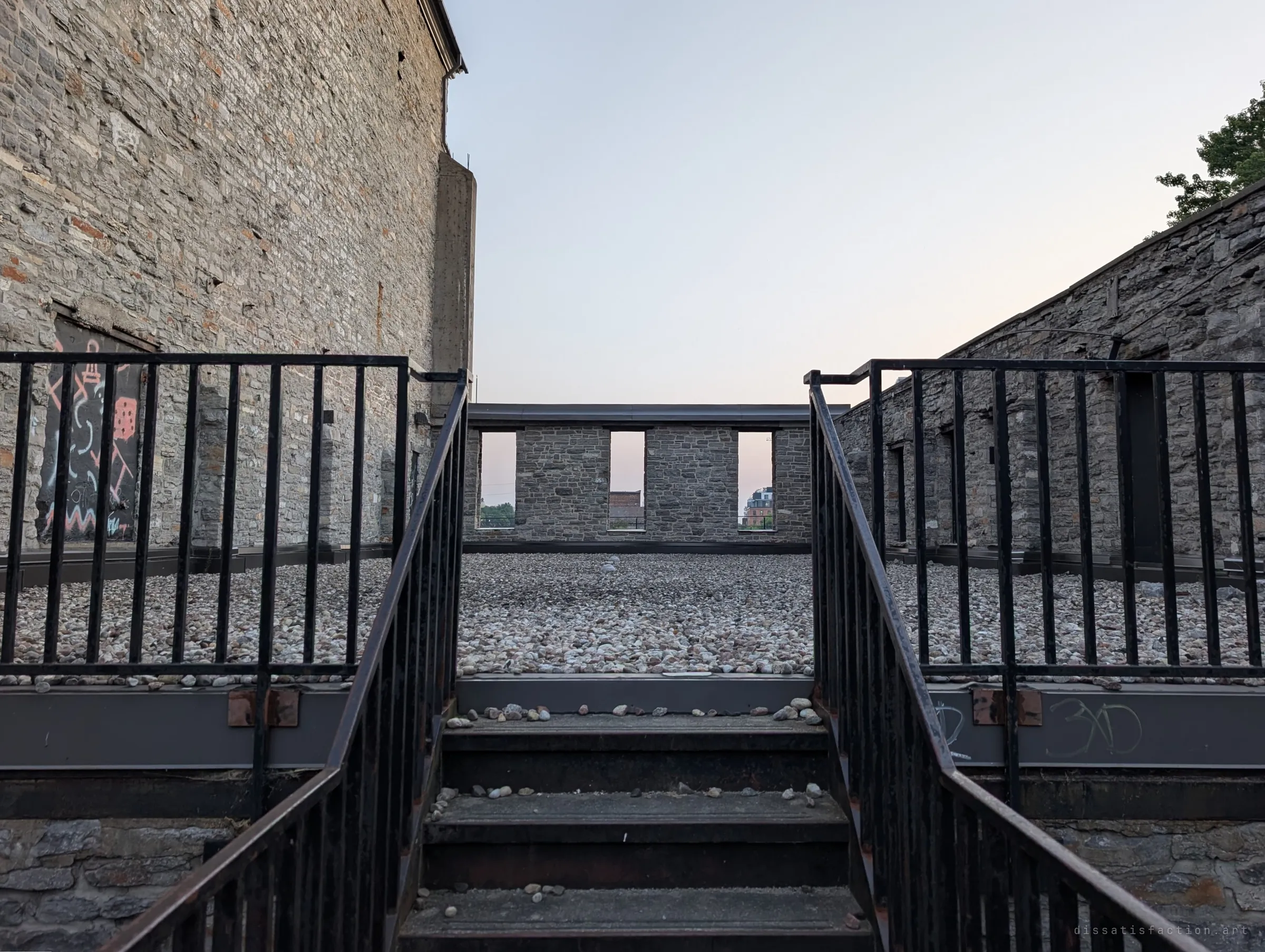A steel stair with black railings climbs onto a gravel-covered roof, where a freestanding stone parapet with three window openings stands against the evening sky. The roof becomes an open terrace inside the ruin.