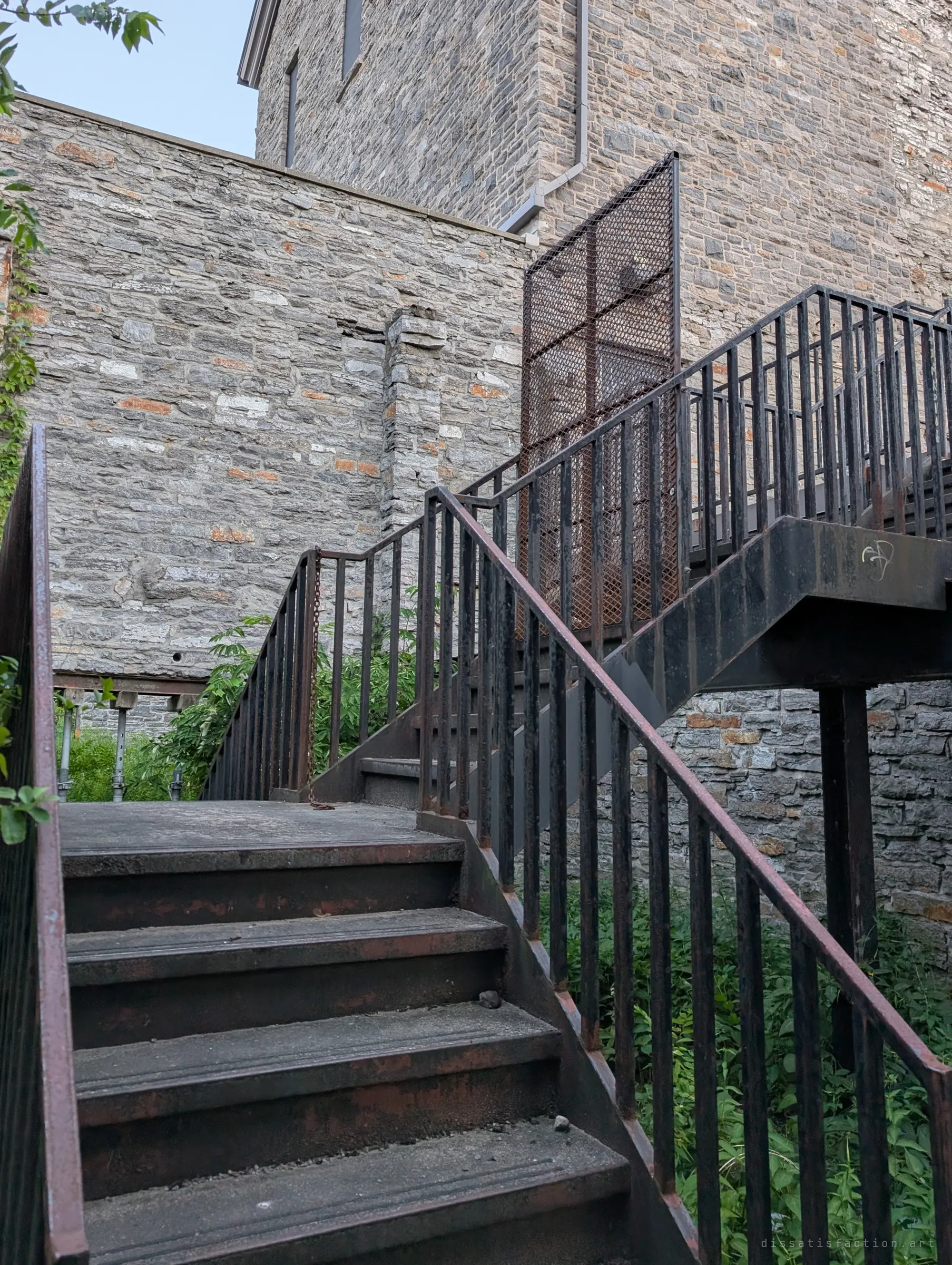 A rusted steel stair climbs past a caged landing against the rough limestone walls of the mill, weeds pushing through below. The bolted-on steel shows how the ruin was made walkable again.
