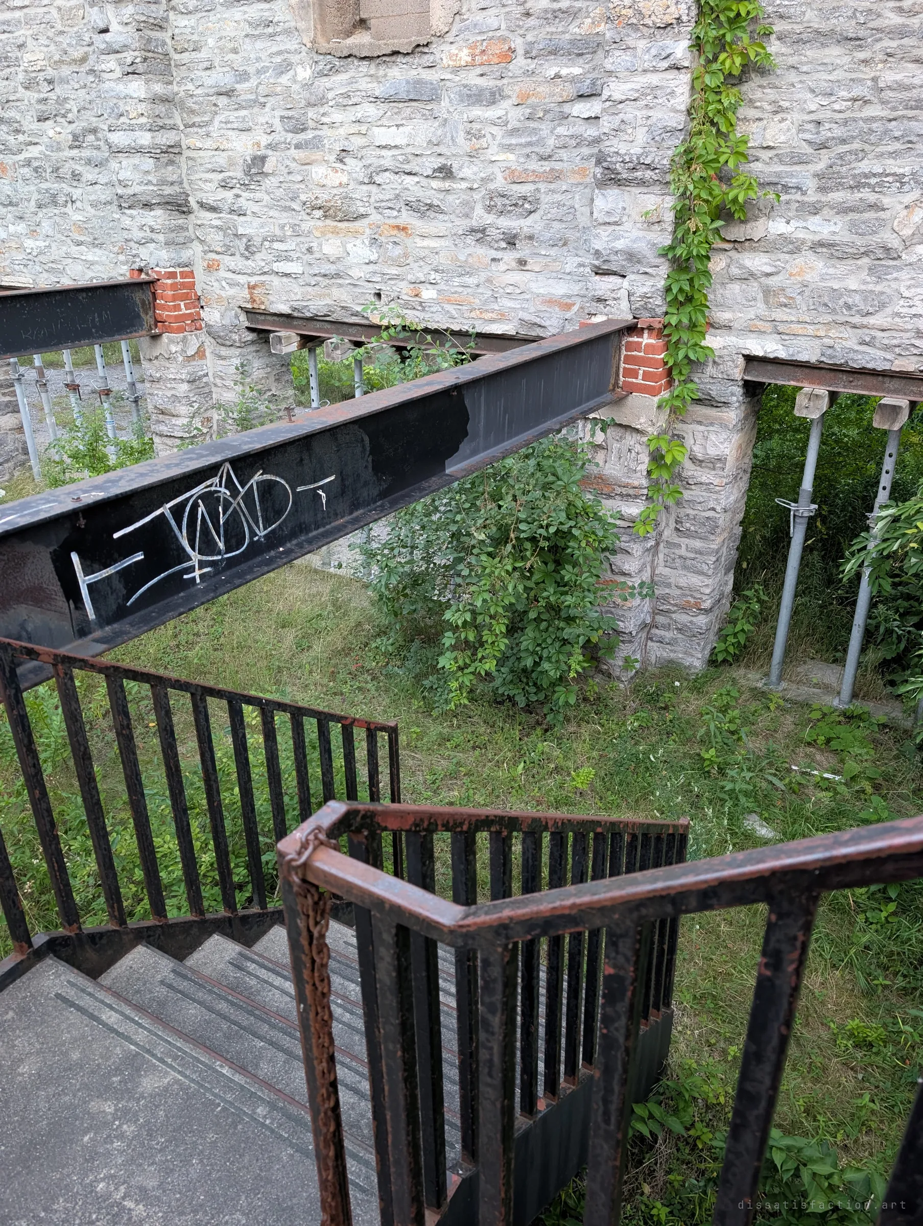 Looking down a switchback steel stair past a tagged I-beam into a grassy courtyard, where galvanised props hold up openings in the stone wall. Virginia creeper climbs the masonry between patched red brick.