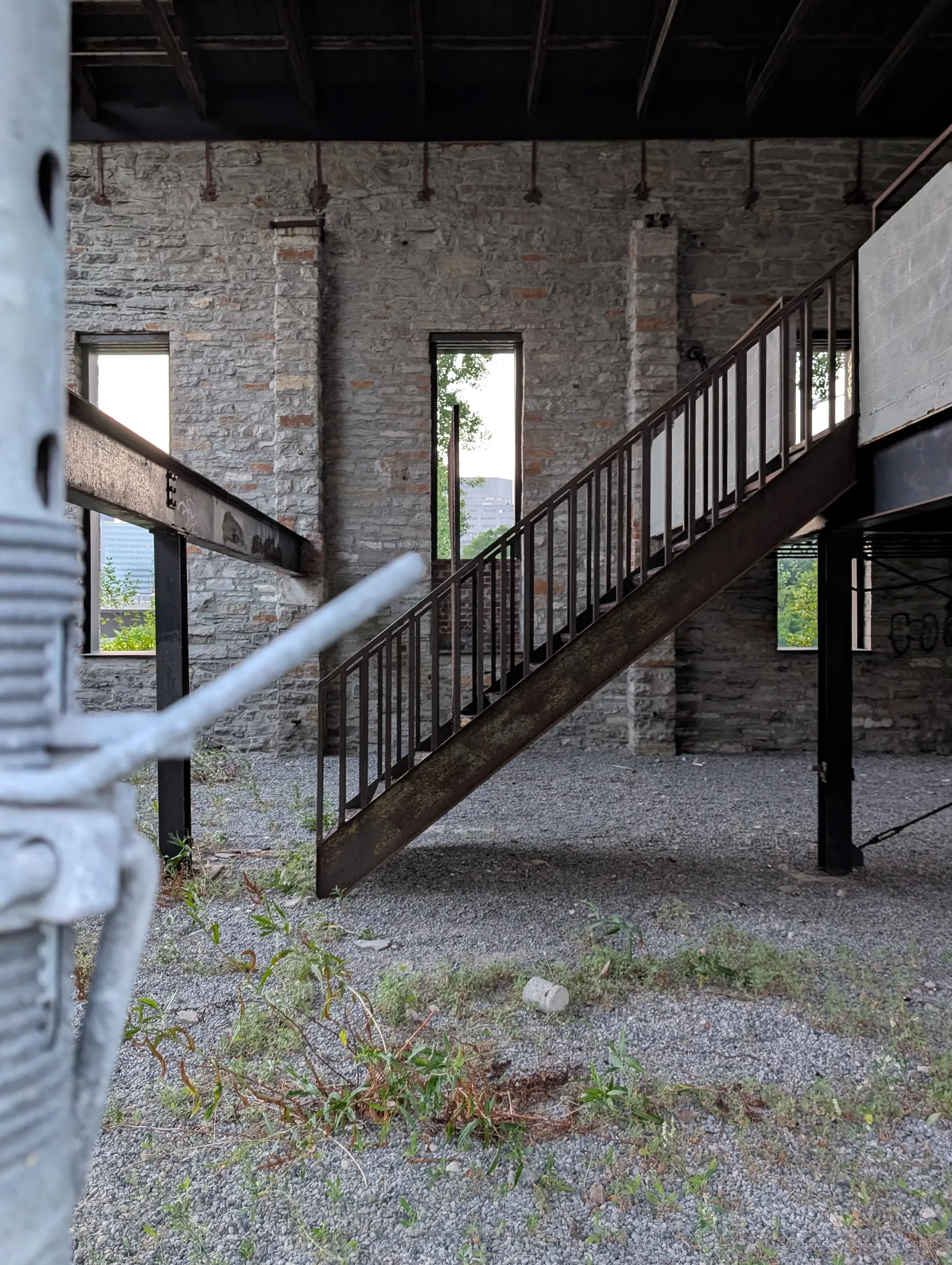 Inside the roofless shell a lone steel stair rises to a mezzanine across a gravel floor, framed by an out-of-focus shoring prop in the foreground. Narrow windows in the stone wall let daylight and greenery in.