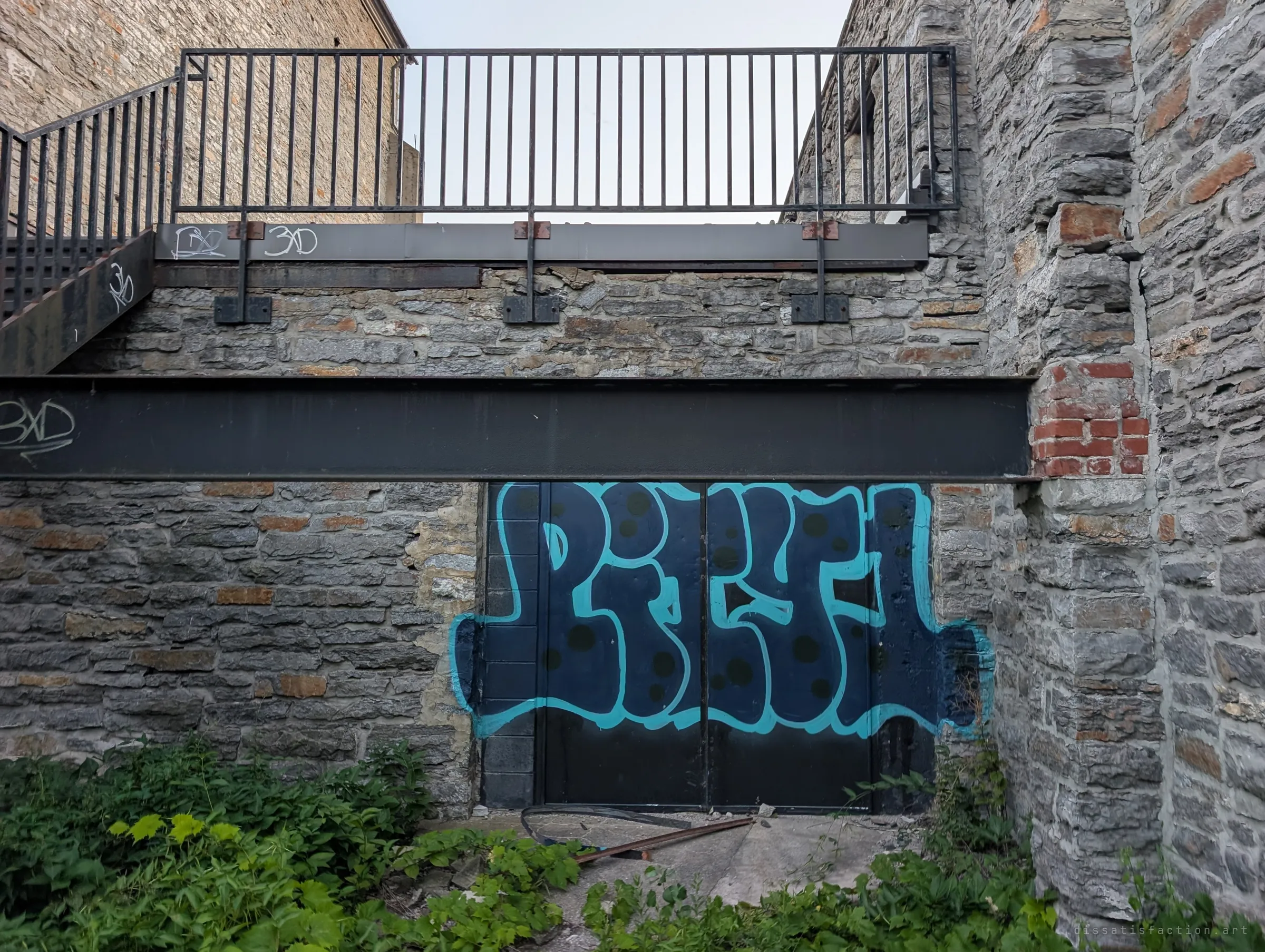 A big blue graffiti piece covers a steel door beneath a heavy black beam, with a railed walkway overhead and stone walls on either side. Tags repeat along the steel, marks of everyone who came before.
