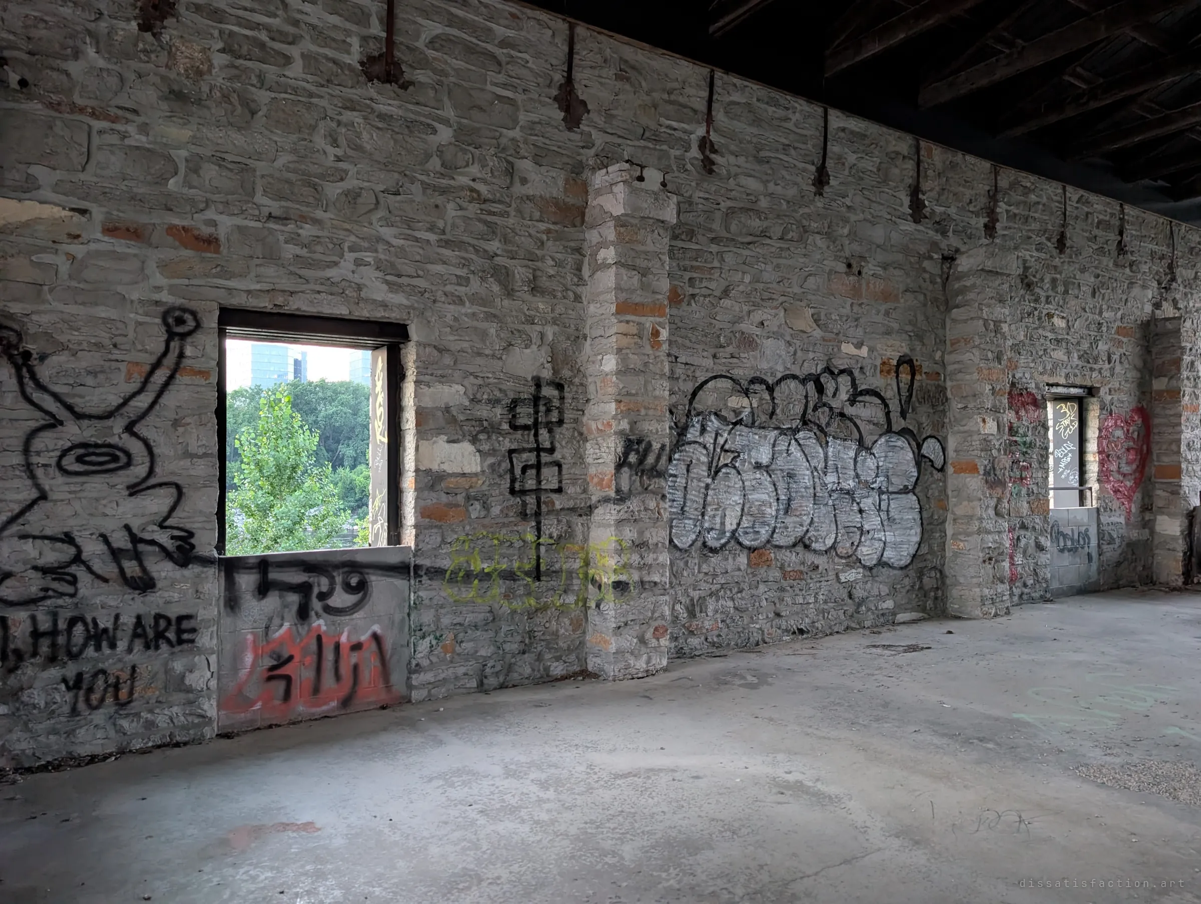 An upper room of bare stone and concrete floor, its walls layered with graffiti including a cartoon figure asking how you are. Rusted rods hang from the dark timber ceiling.