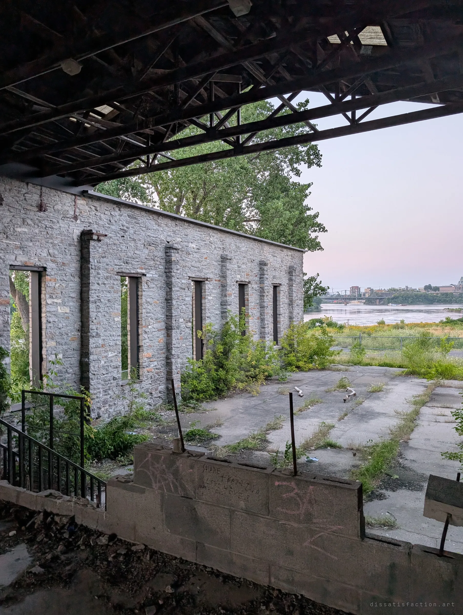 From under a skeletal roof truss, a roofless stone wall of tall empty windows stands beside a cracked concrete yard going green. Pink dusk light sits over open water beyond.