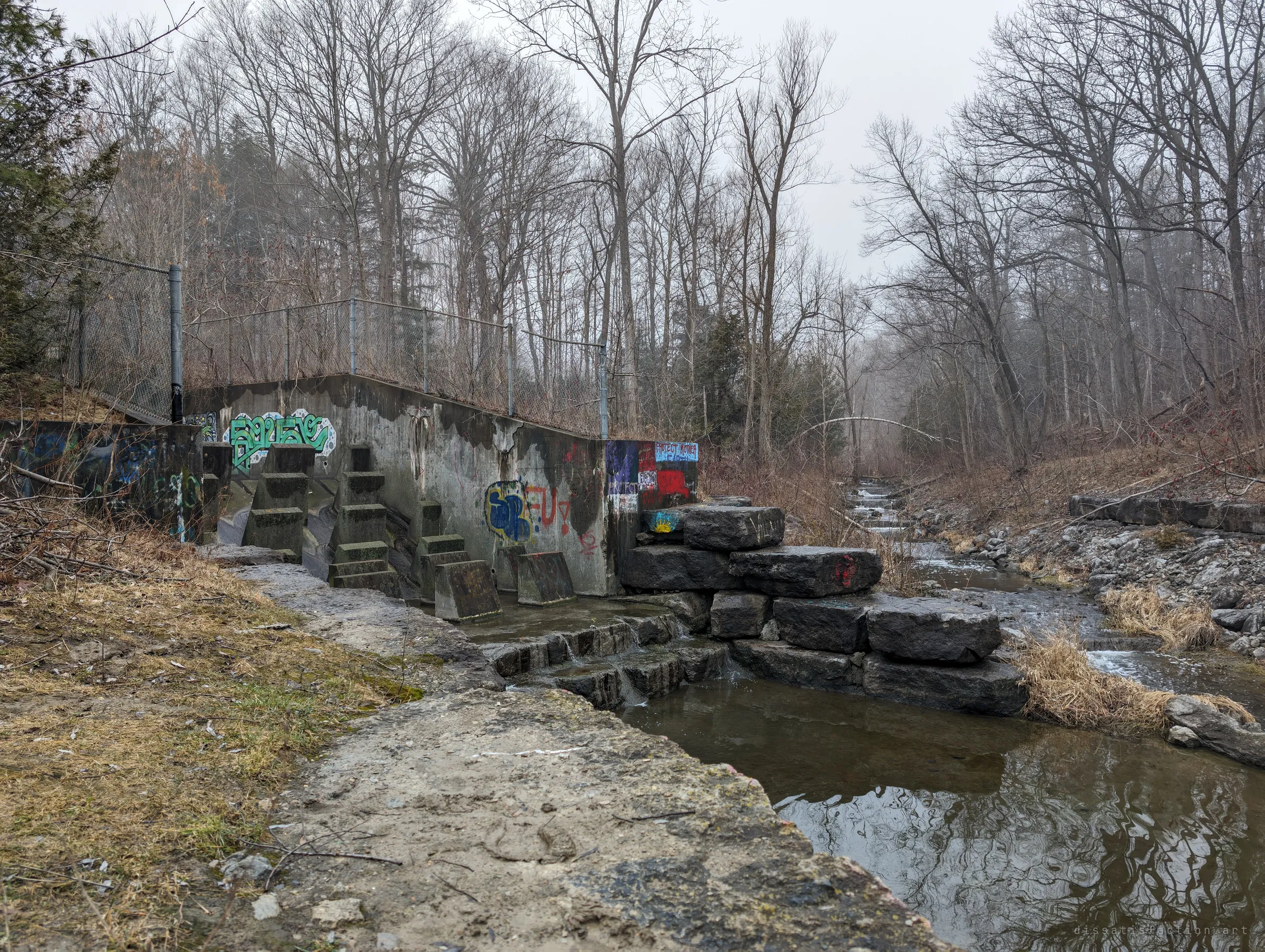 A graffiti-covered concrete drop structure sits in a misty, leafless creek valley, water spilling over stone blocks into a still pool. It sets the scene: the outfall where the drain meets daylight.