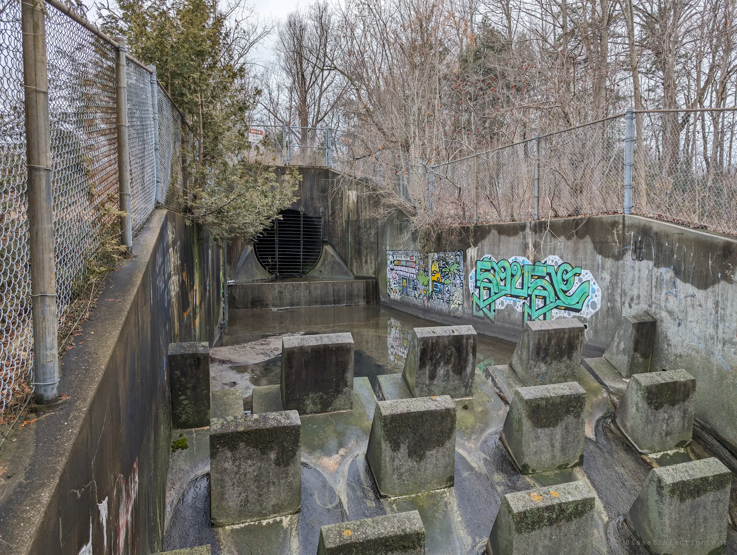 Behind the concrete baffle blocks, a barred circular outfall opens into a flooded basin lined with colourful murals and chain-link fencing. The first look at the drain mouth itself.