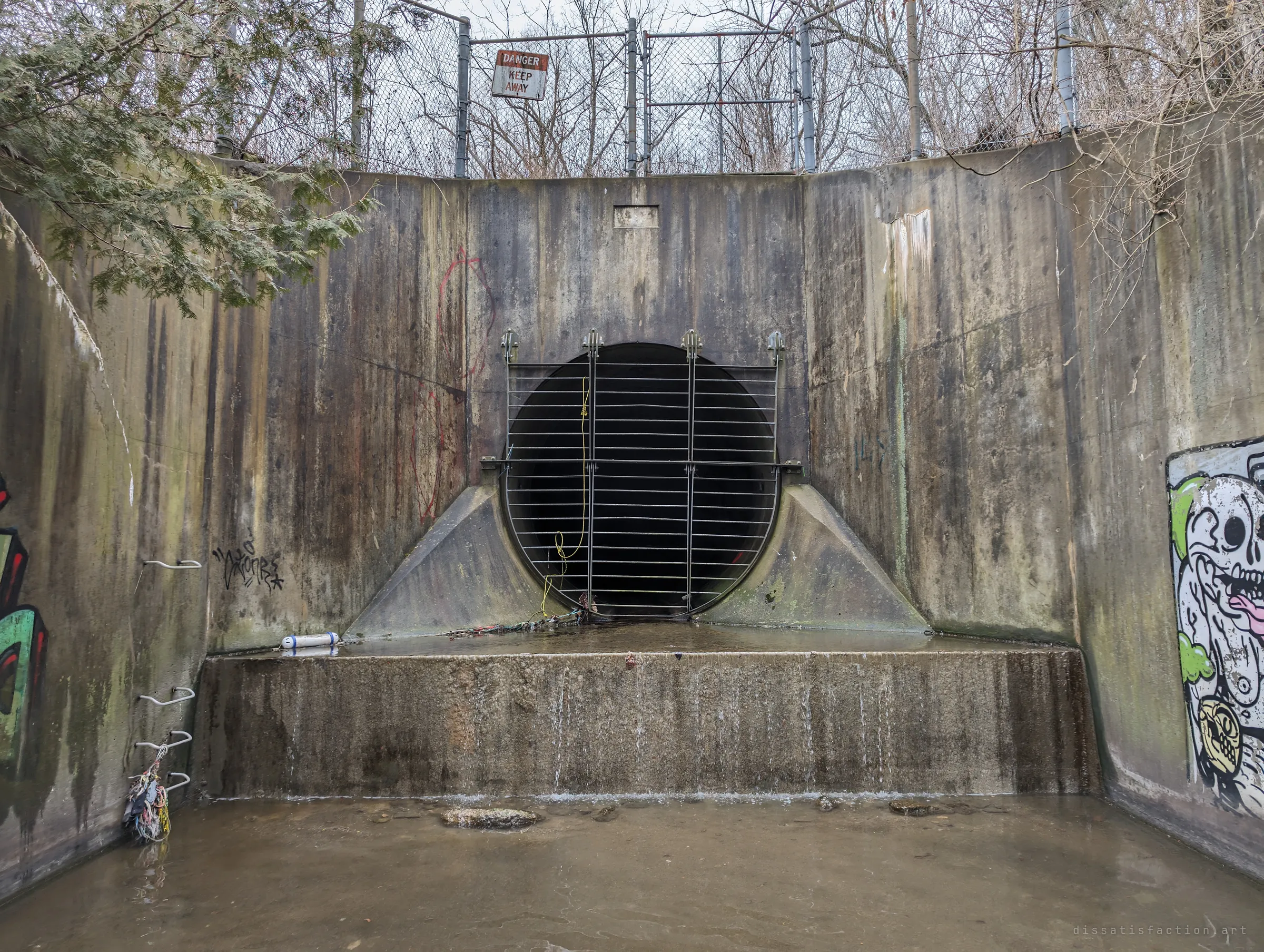 A round storm drain outfall sealed by a hinged metal grate, set in a streaked concrete headwall with a generic danger sign on the fence above. Rungs on the side wall and a shallow pool at its feet complete the approach.