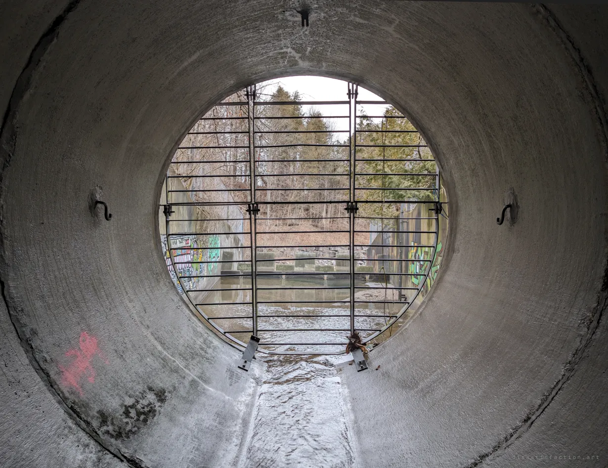 From just inside the pipe, the circular grate frames the basin outside with its murals and baffle blocks. The view back to daylight once past the bars.