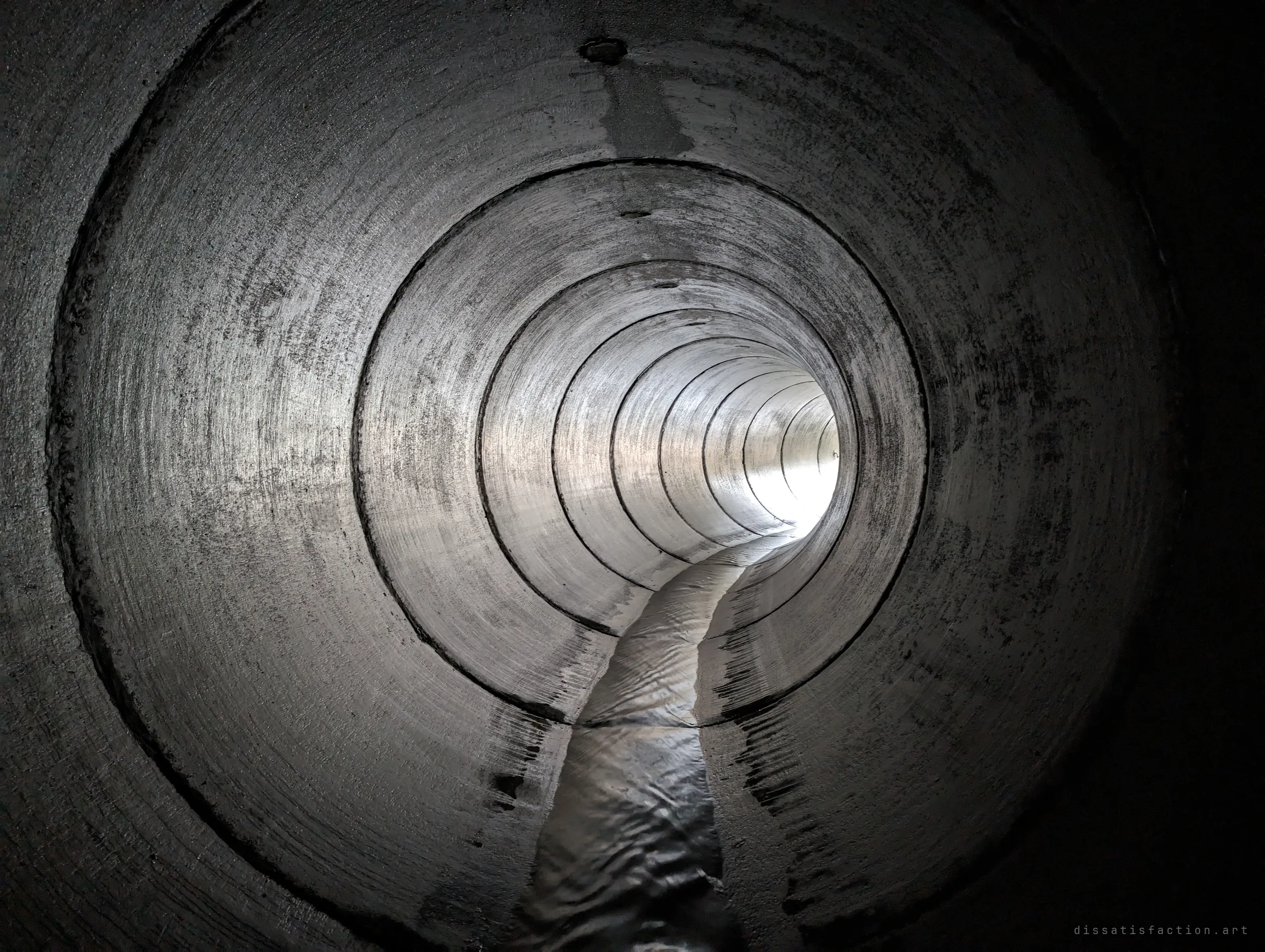 A smooth concrete pipe recedes in stacked rings toward a bright opening, a thin stream of water running along its floor. The simple repeating geometry of the drain interior.