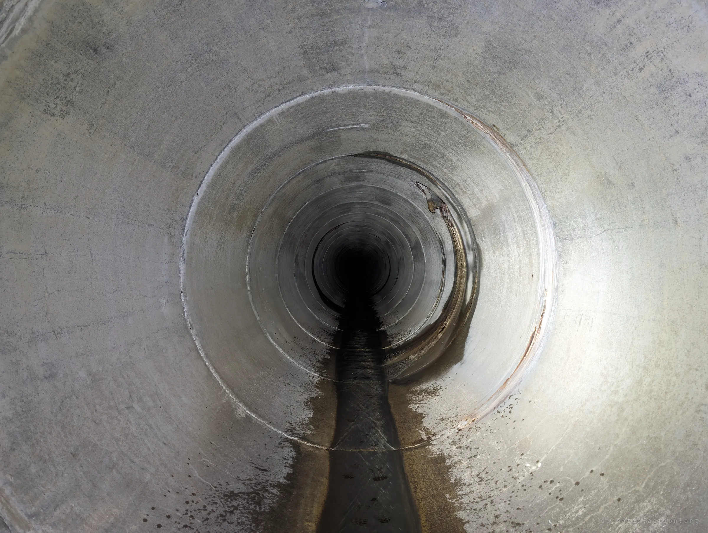 Looking into a narrower pipe step-down, concentric concrete rings shrink into total darkness with a dark trickle of water along the invert. Marks where the drain continues on out of reach of daylight.