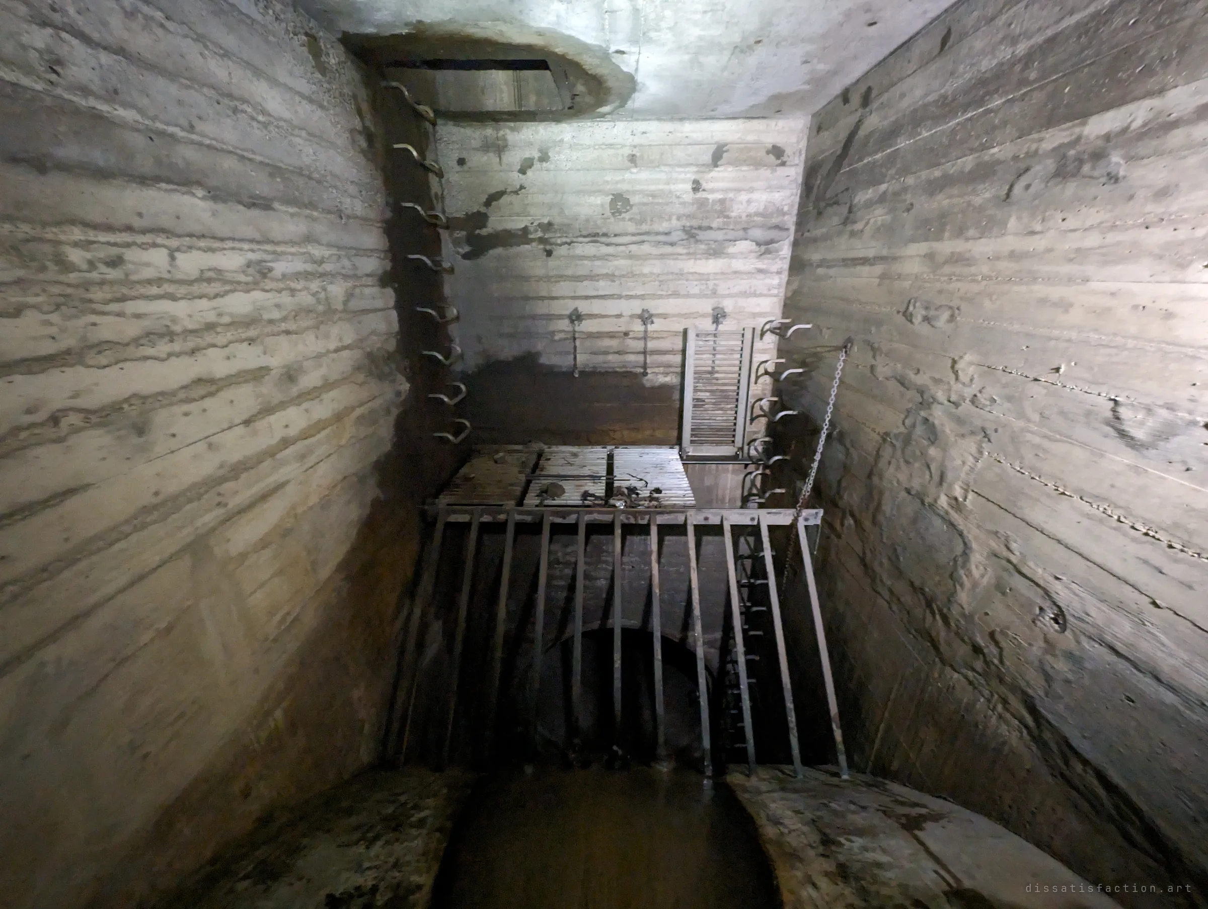 A tall board-marked concrete chamber with step irons climbing to a manhole above, a steel trash screen and grated platform guarding the pipe below. It shows the access shaft and debris screen at the heart of the system.