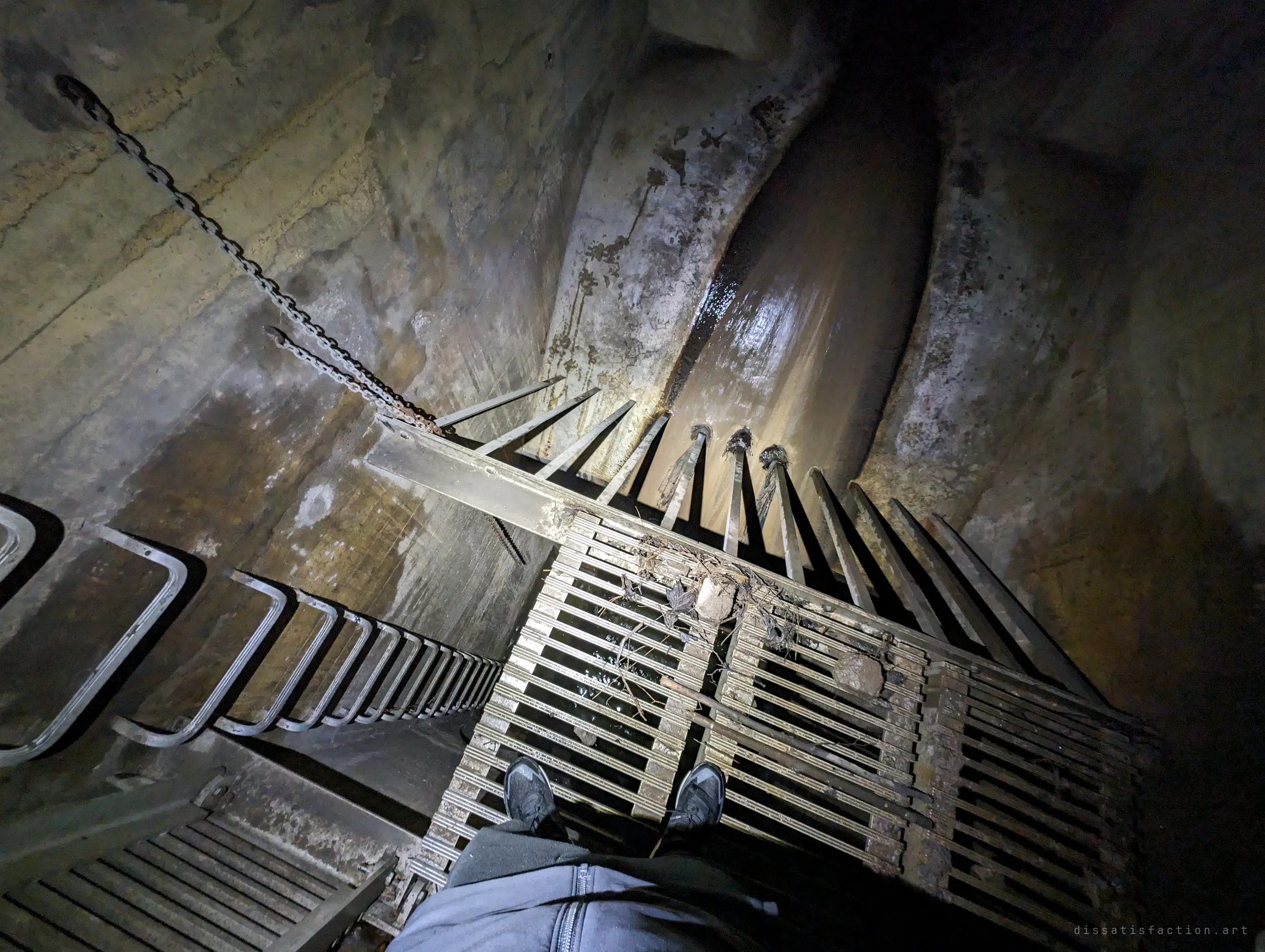 Looking straight down from a grated steel platform, the photographer's shoes rest above a trash rack hung with debris, while a chain and ladder drop into the brown water channel. It captures the vertigo of standing on the screen over the flow.