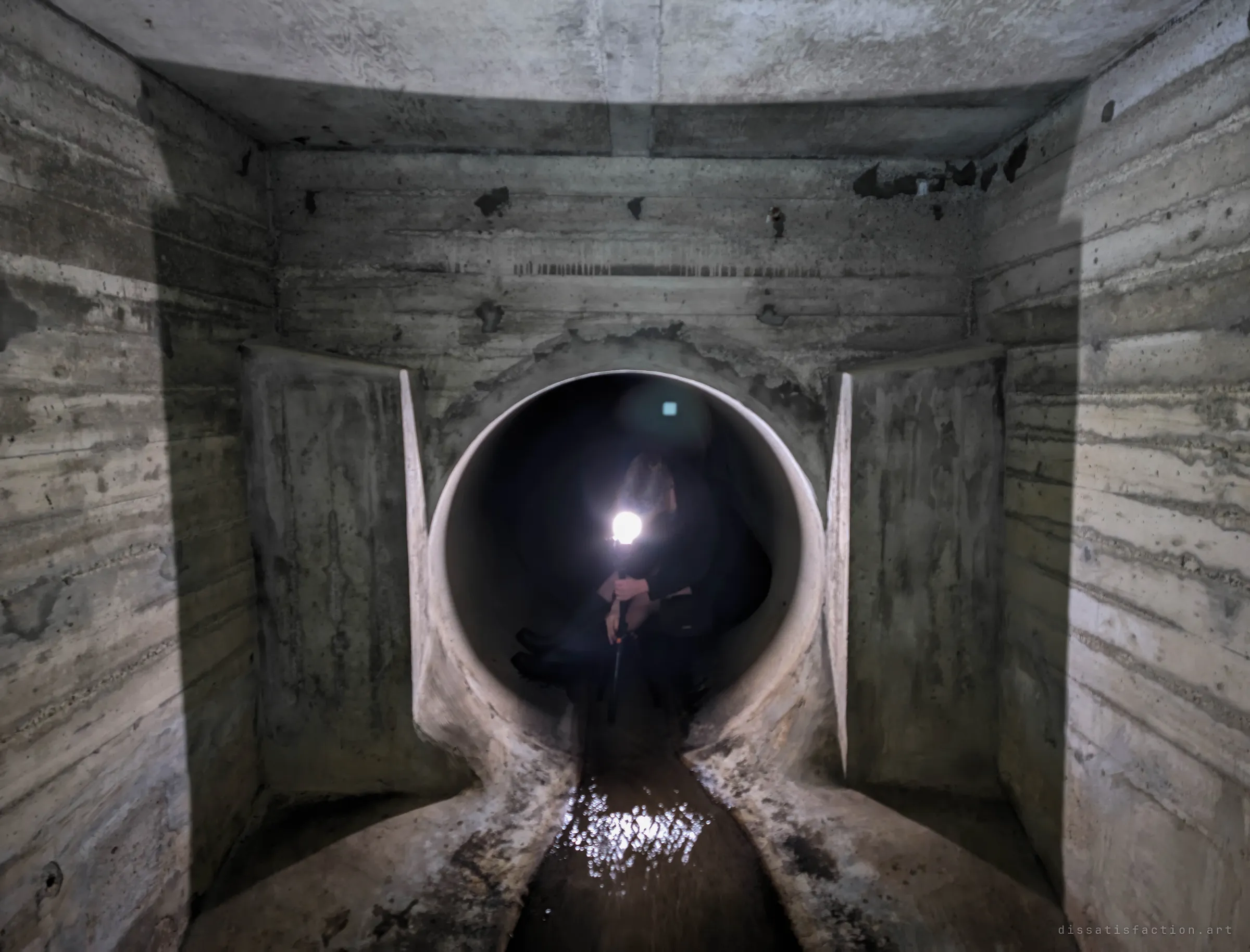 A crouching figure holds a bright torch inside a round pipe where it enters a boxy concrete chamber, the beam glinting off the water channel. The person gives scale to the pipe and the chamber around it.