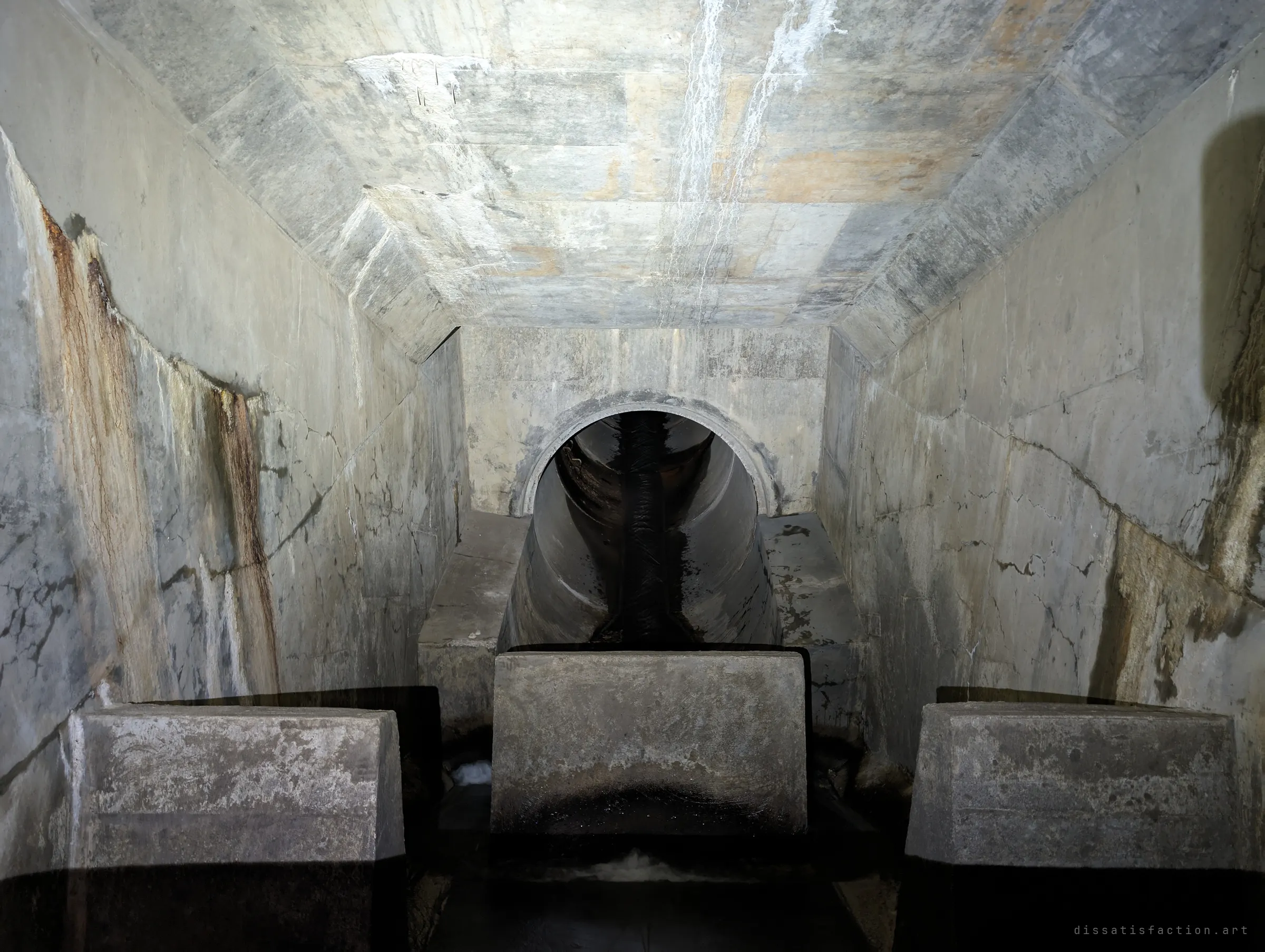 A long concrete chamber with a chamfered ceiling and rust-streaked walls leads to a single round pipe, with three heavy concrete baffle blocks standing in the dark water in front. The blocks slow the flow before it leaves the chamber.