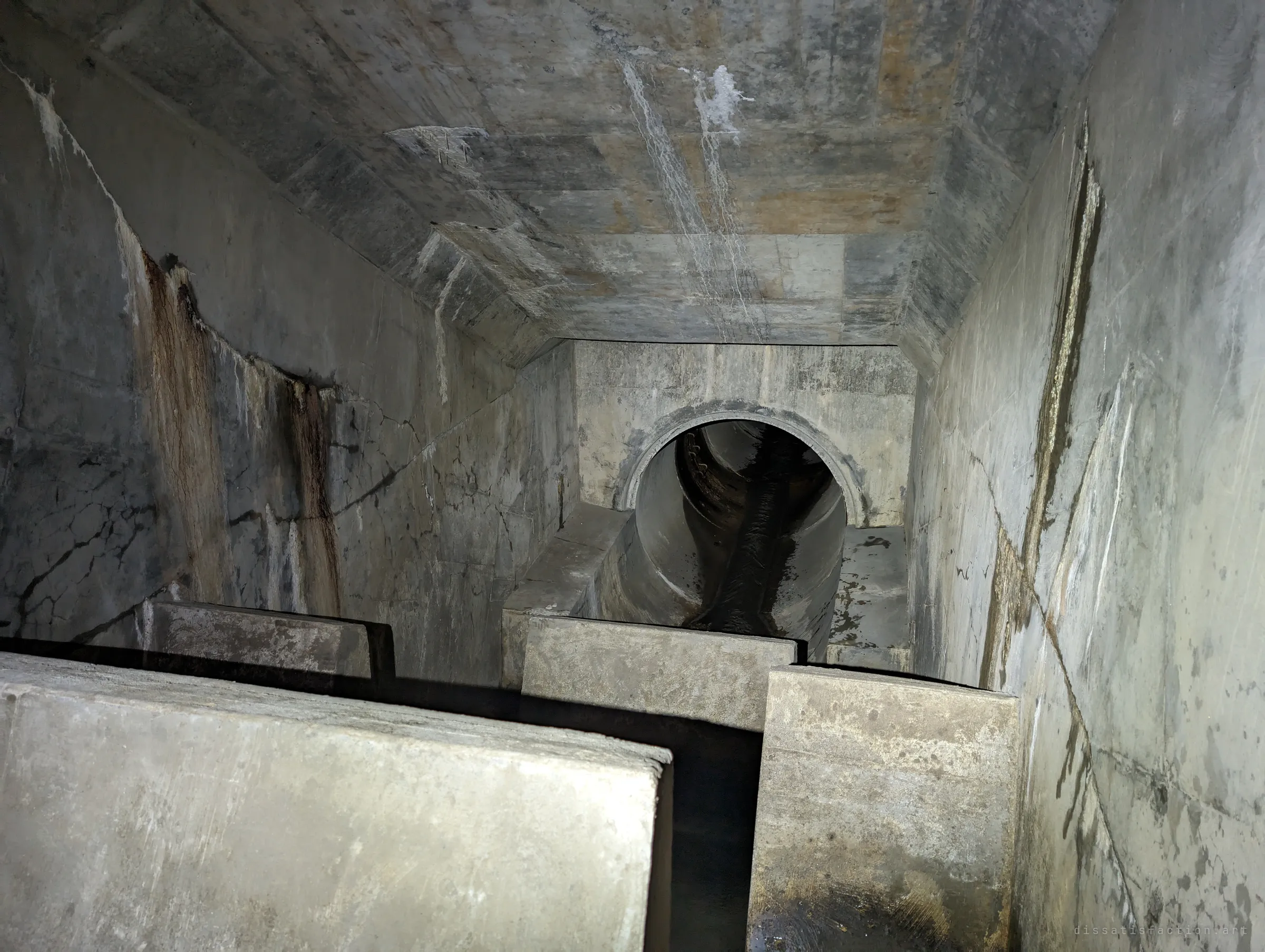Seen from above, concrete baffle blocks crowd the foreground of a rust-streaked chamber that narrows to a round outlet pipe. The steep angle shows how deep the energy-dissipating blocks sit.
