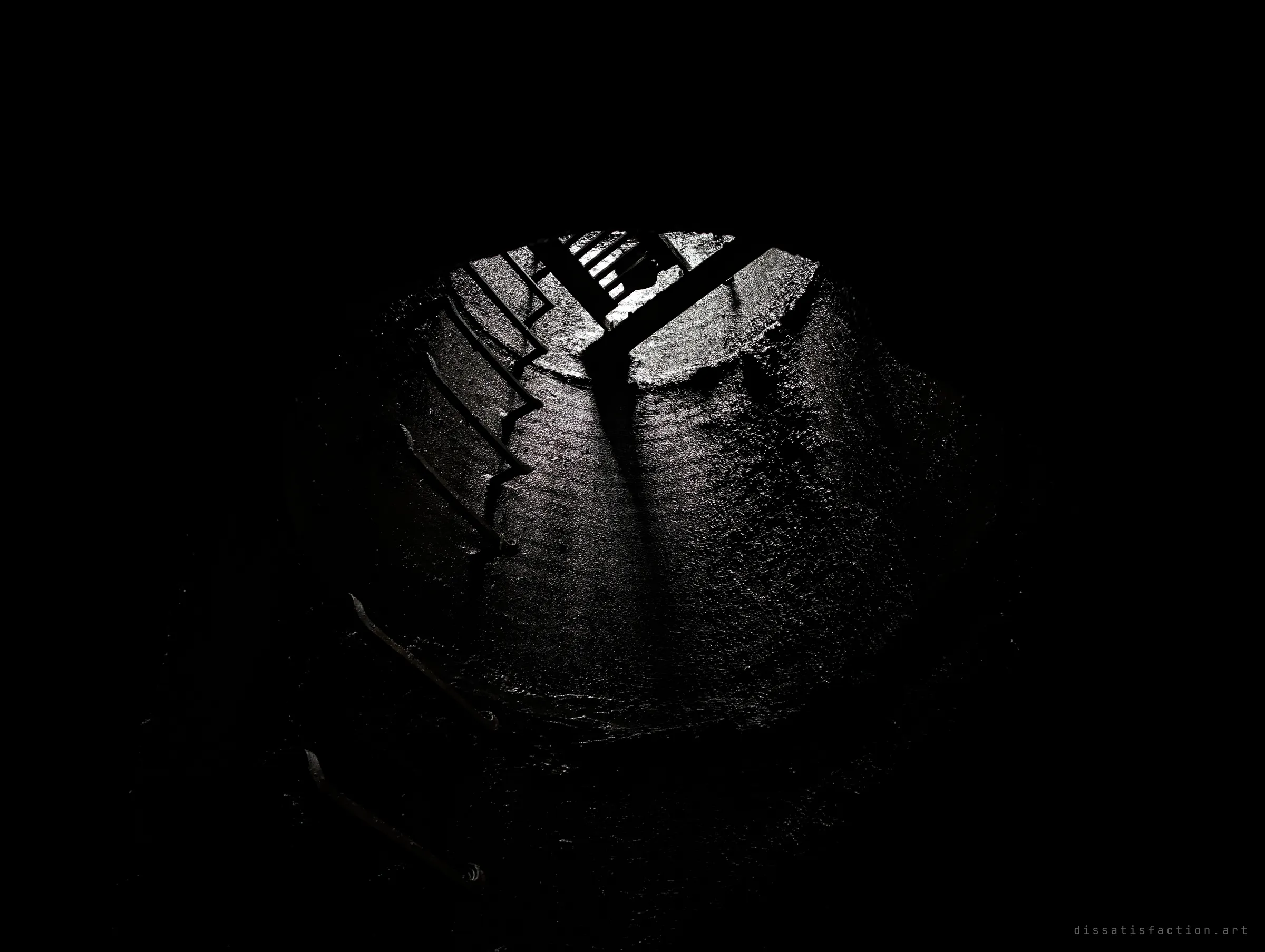 Almost total darkness frames a round shaft lit from above, where a grated hatch and step irons cast long shadows down the wet concrete. It shows the only way in and out, seen from the bottom.