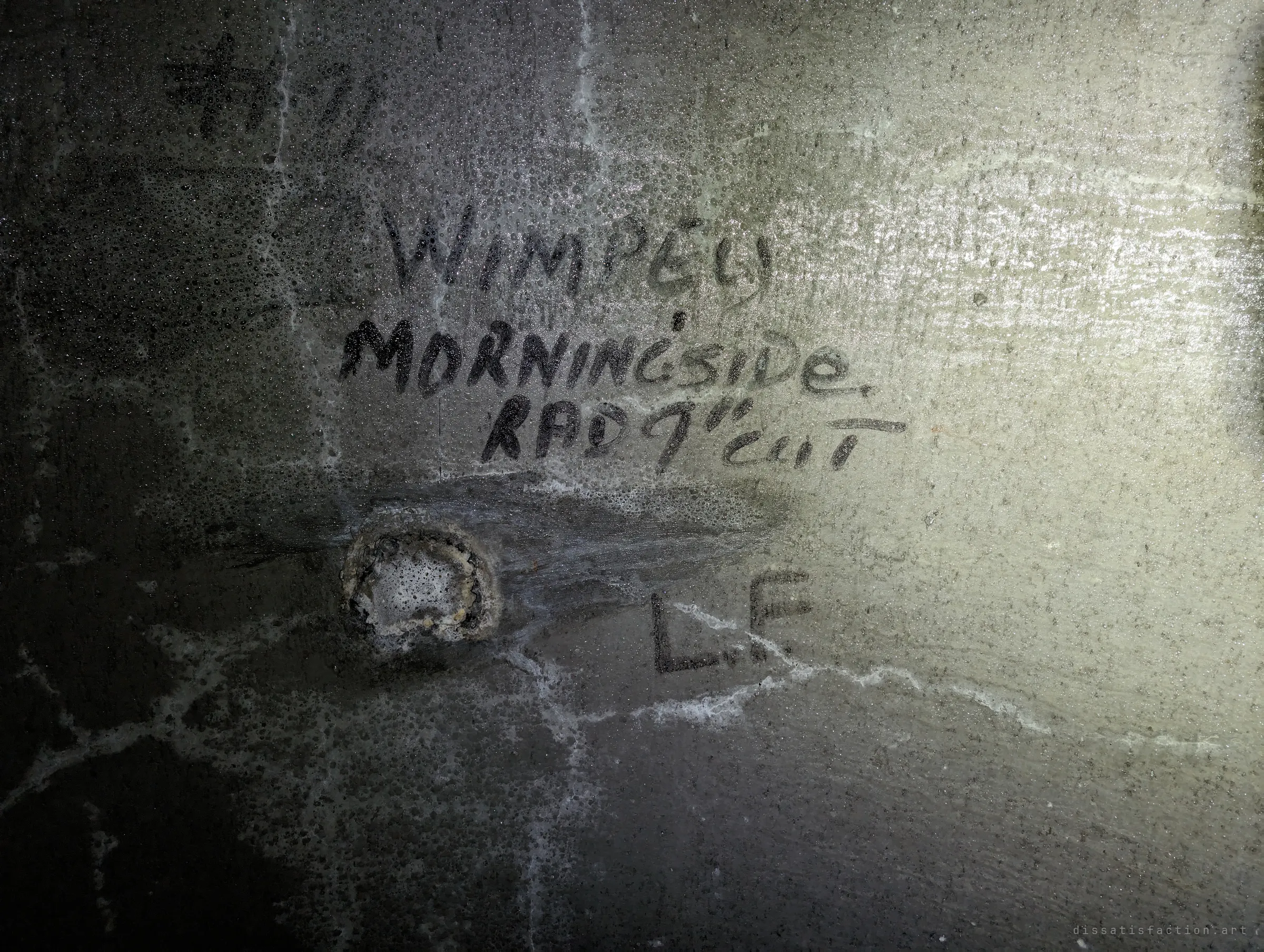 Old handwritten builder's markings in black paint on a damp concrete wall, beside a plugged tie hole and a pair of initials. A trace of the crew who cast this chamber decades ago.