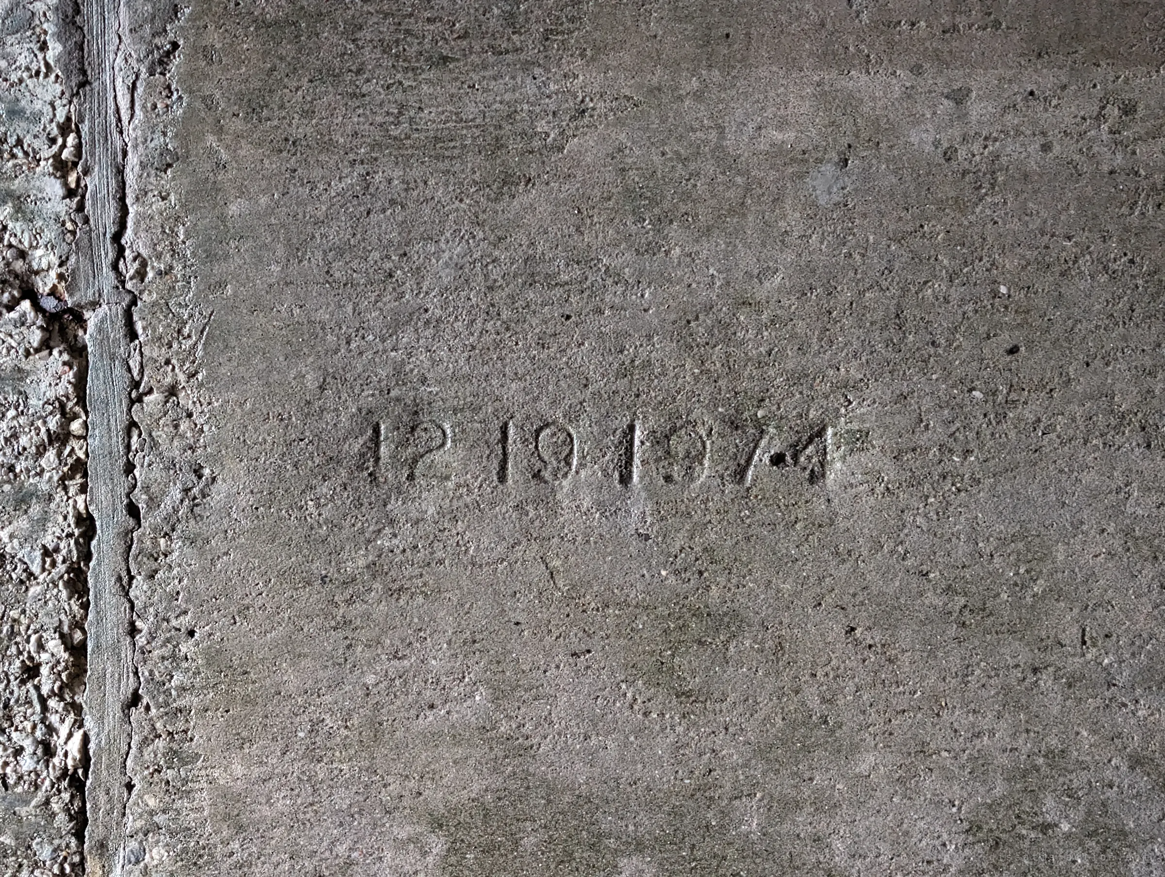 A construction date stamped into smooth concrete beside a rough aggregate joint. It dates the drain to the mid-1970s.