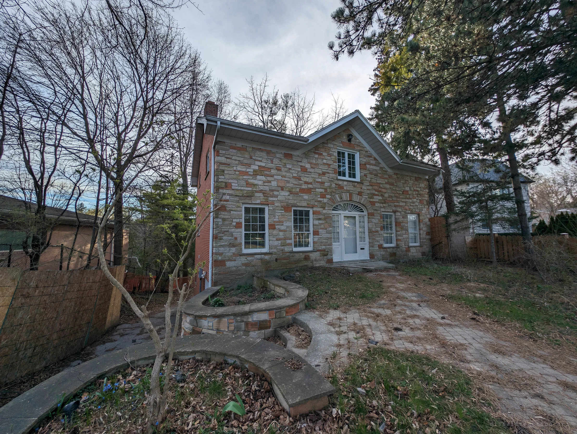 A stone-faced two-storey house with a fanlight over the front door sits behind curved garden walls, bare trees and a leaf-strewn paver path. The establishing shot of an empty home going quiet.