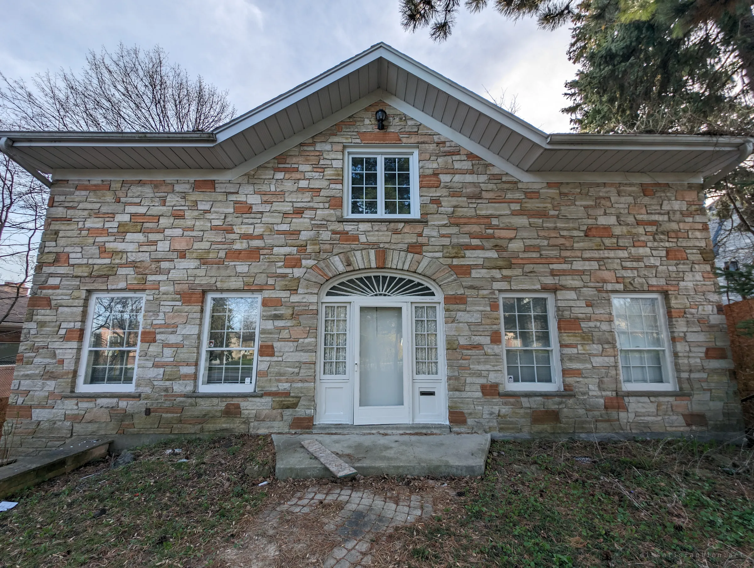 The front of the house head-on: patchwork stone walls, white sash windows and an arched fanlight over the door, with a loose stone slab lying on the front step.