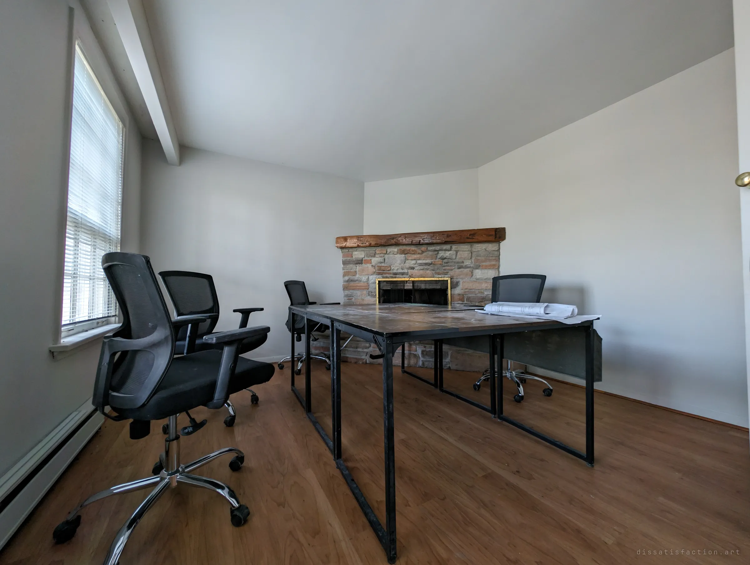 Office chairs circle a pair of metal-framed desks in a front room with a stone corner fireplace; rolled plans lie on the table as if work stopped mid-meeting.