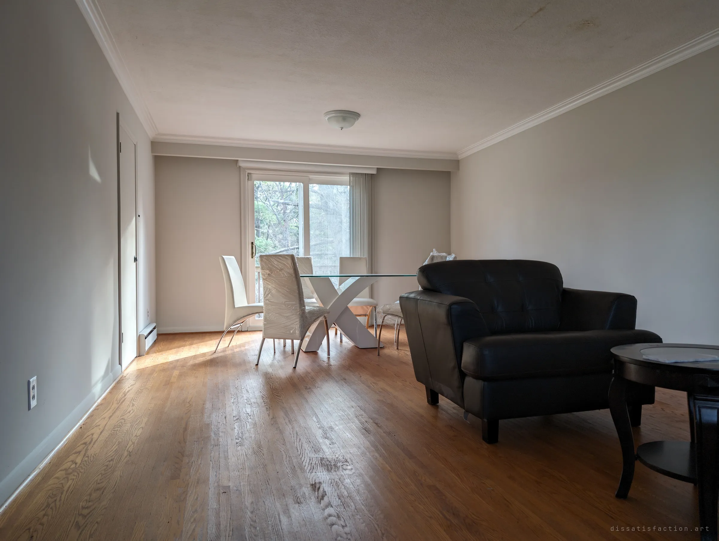 A long living room with oak floors, a leather armchair and a glass dining table whose white chairs are still wrapped in plastic, lit by a sliding door to the back.