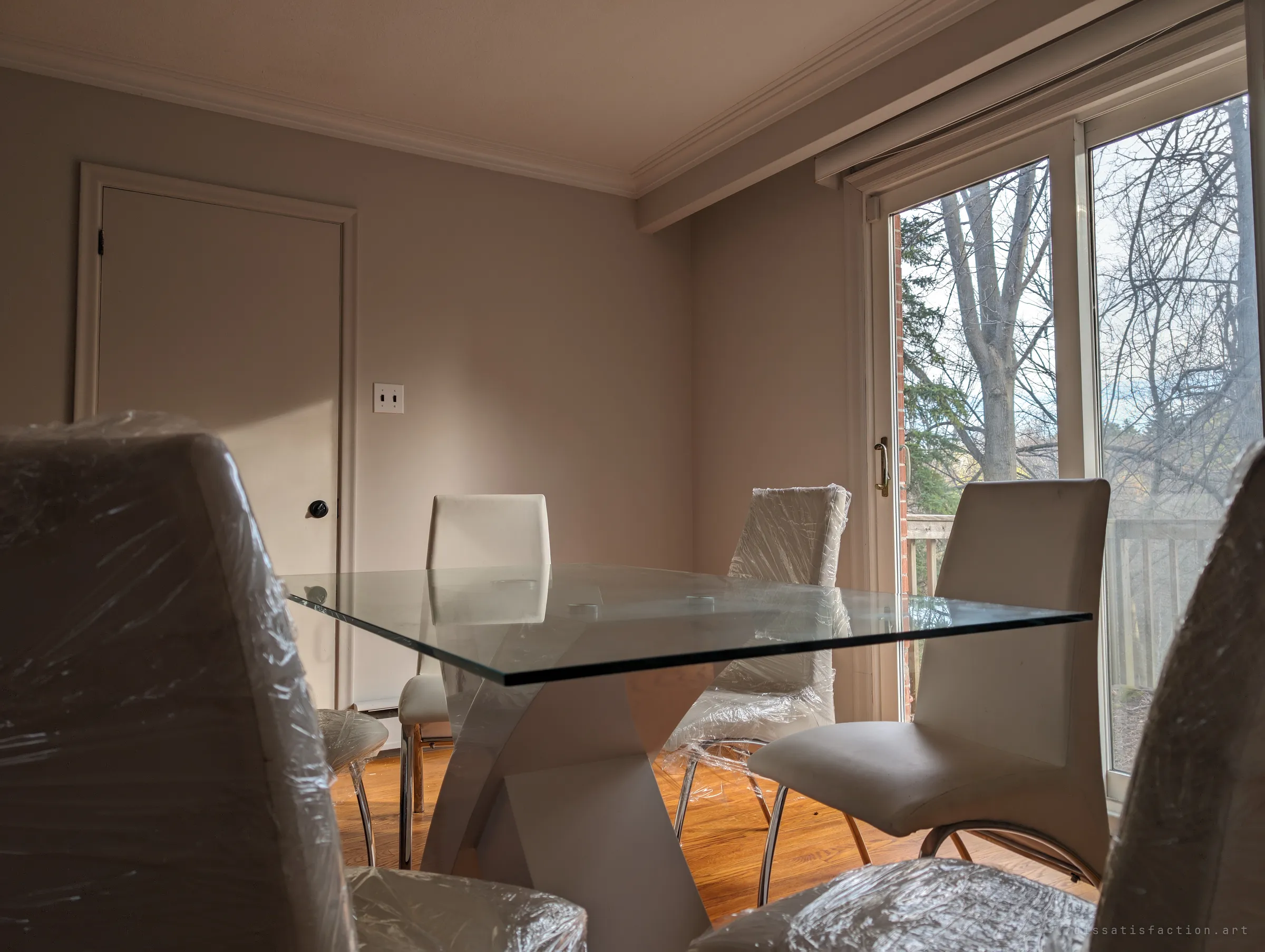 Low view across the glass dining table, its chairs still sealed in shrink-wrap, toward a sliding door onto a deck and bare trees. New furniture that was never used.