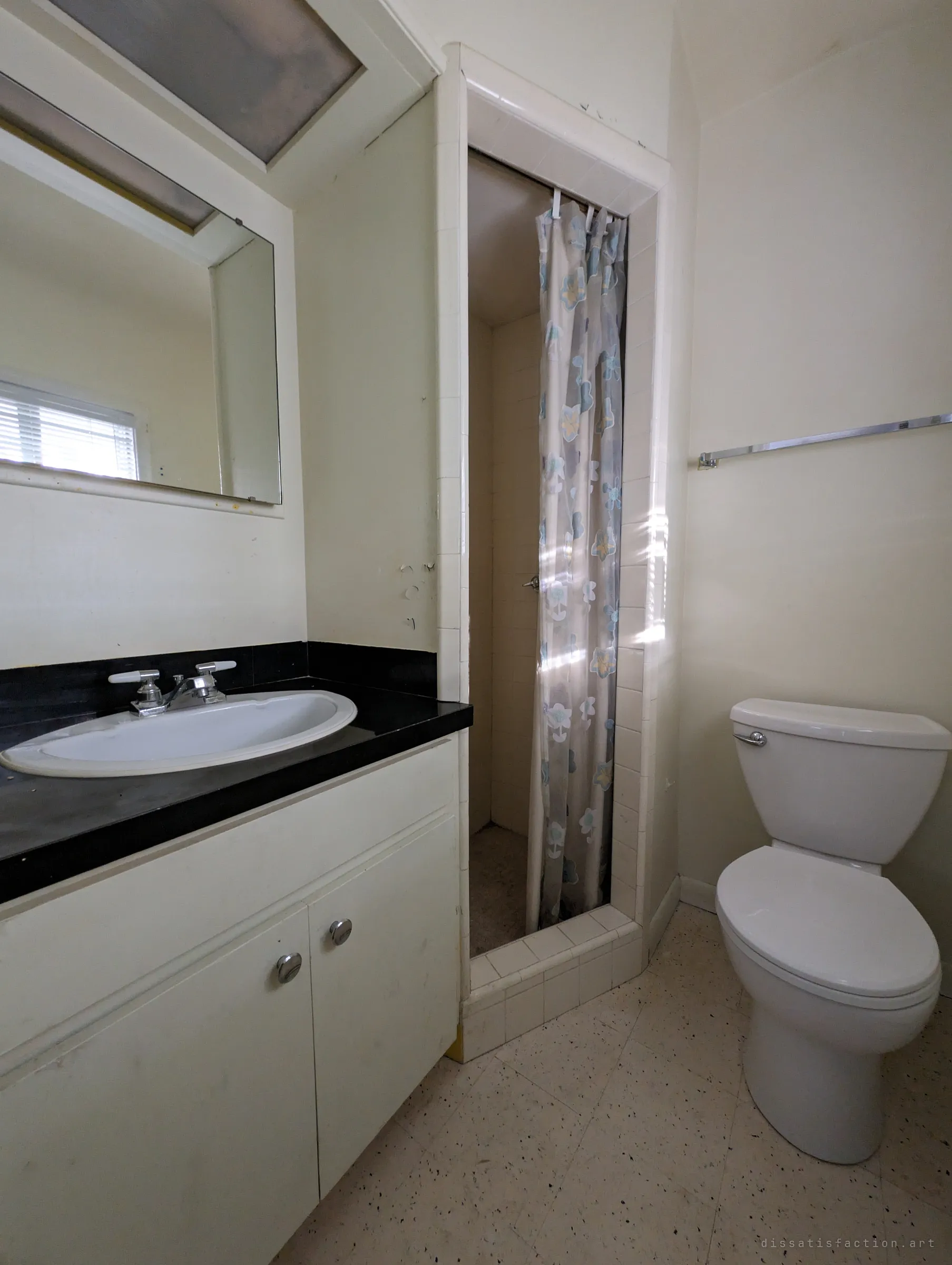 A dated bathroom with a black vanity top, a narrow tiled shower stall behind a floral curtain and speckled floor tiles in soft daylight.