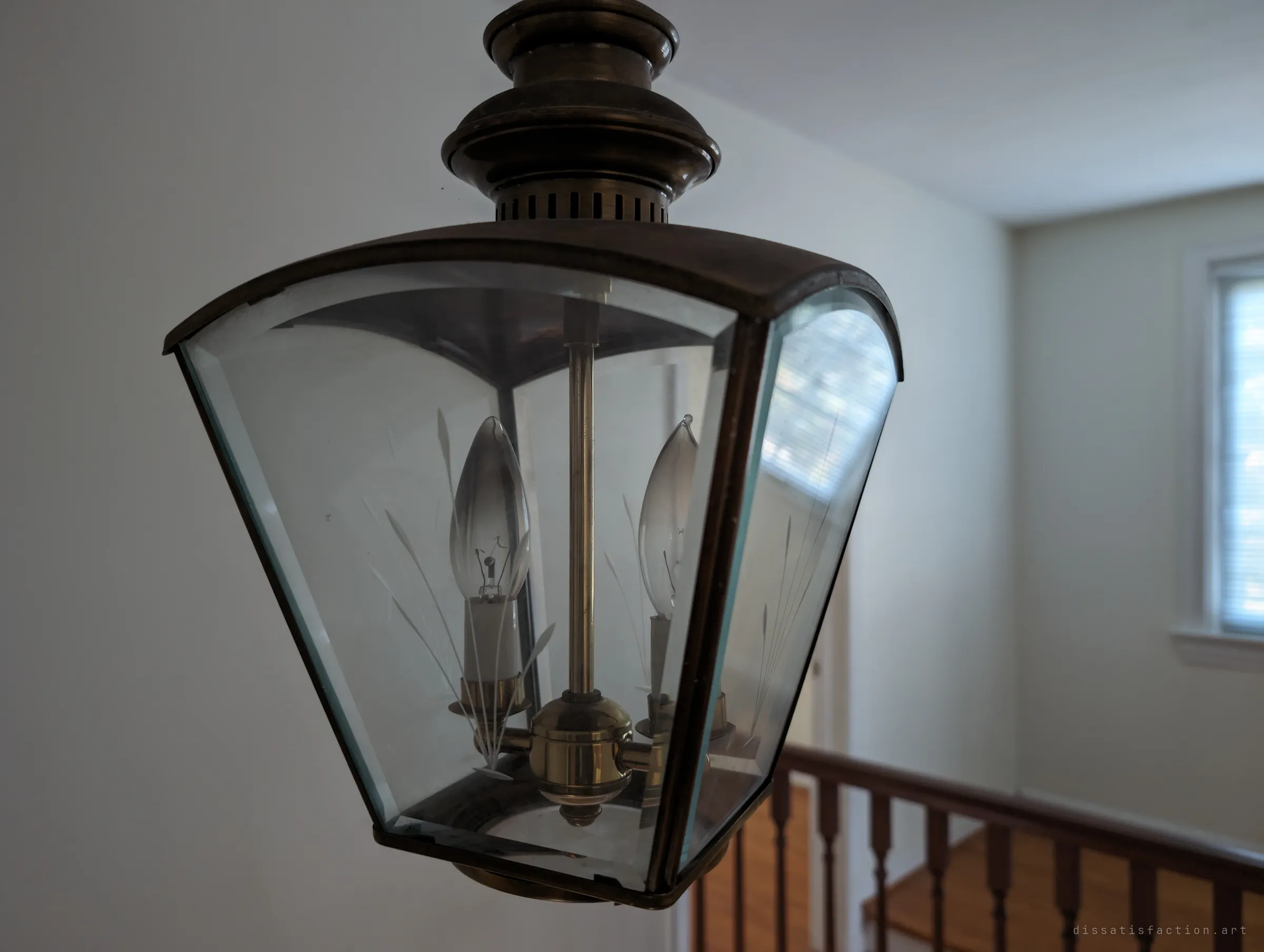 Close-up of the etched-glass hall lantern with its two candle bulbs, the stair railing blurred below. A small period detail left behind in the empty house.